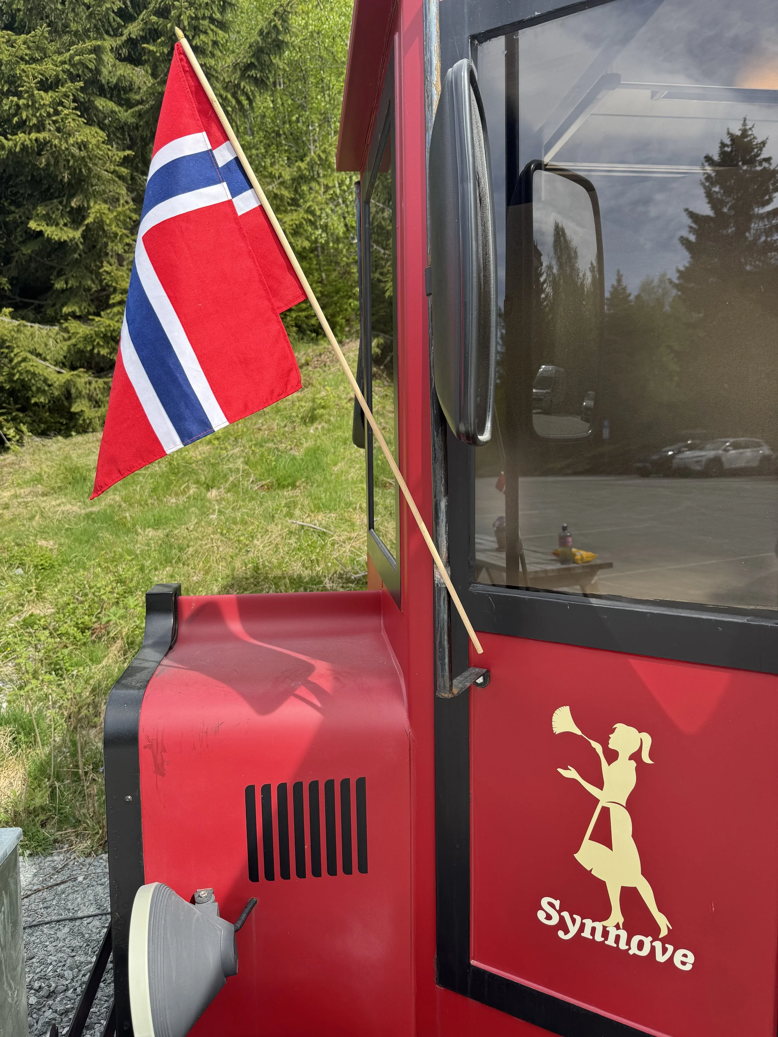 Norsk flagg festet på en liten rødt kjøretøy, med en logo av en kvinne som blåser i en horn og teksten 'Synnøve' på siden, omgitt av grønne trær.