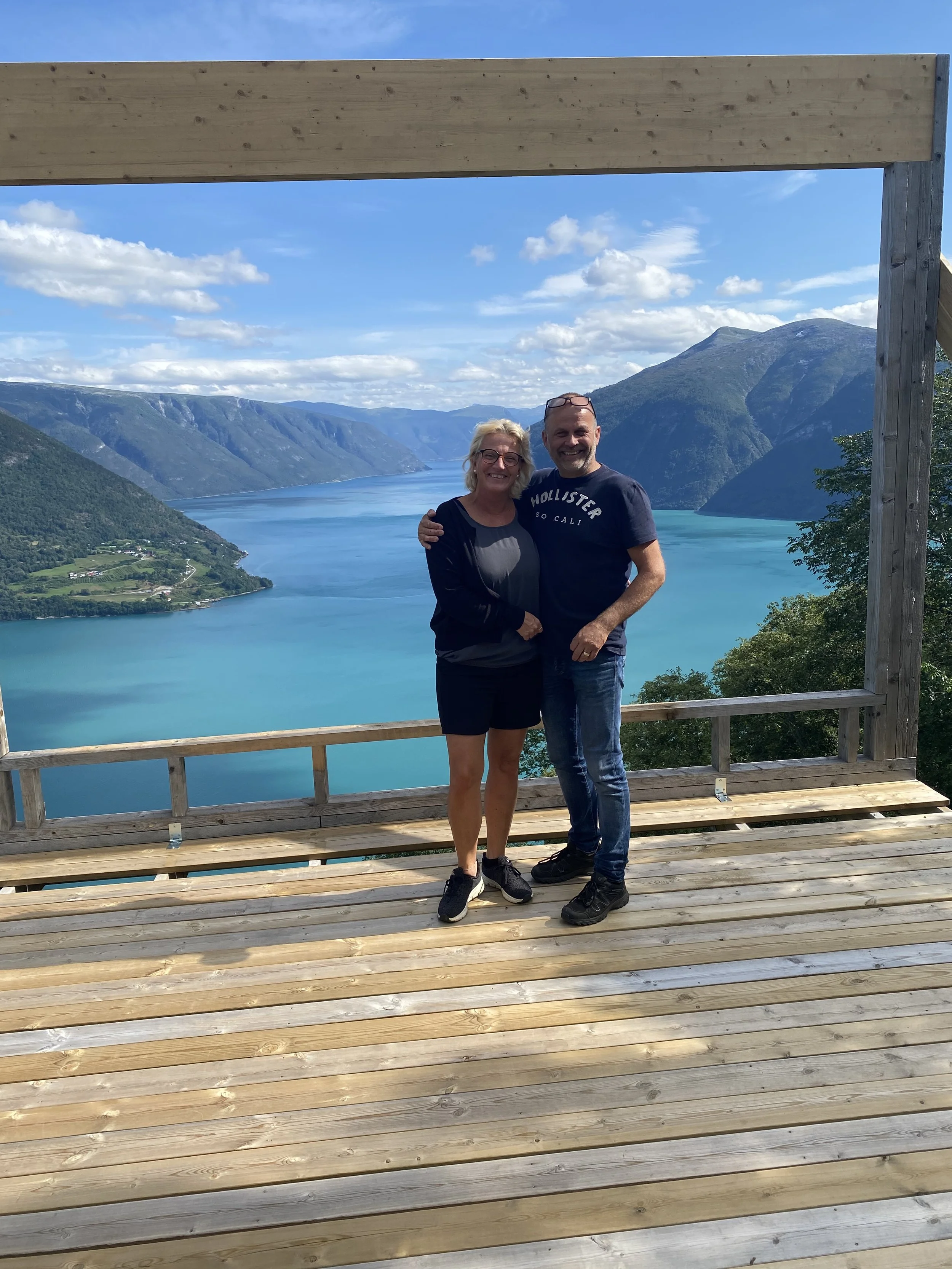 To personer står på en tredekket plattform med utsikt over en fjord omgitt av fjell og en klar blå himmel.