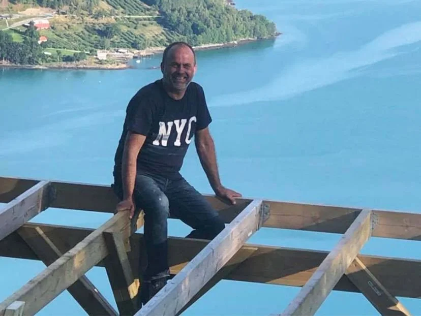 En mann som smiler, sitter på en trekkel over en innsjø med landskap i bakgrunnen.