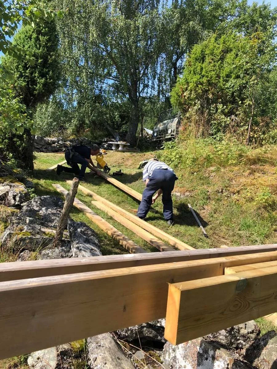 To personer som bygger en trebro i en hage, med tre lange staver og verktøy, omgitt av grønne trær og busker.