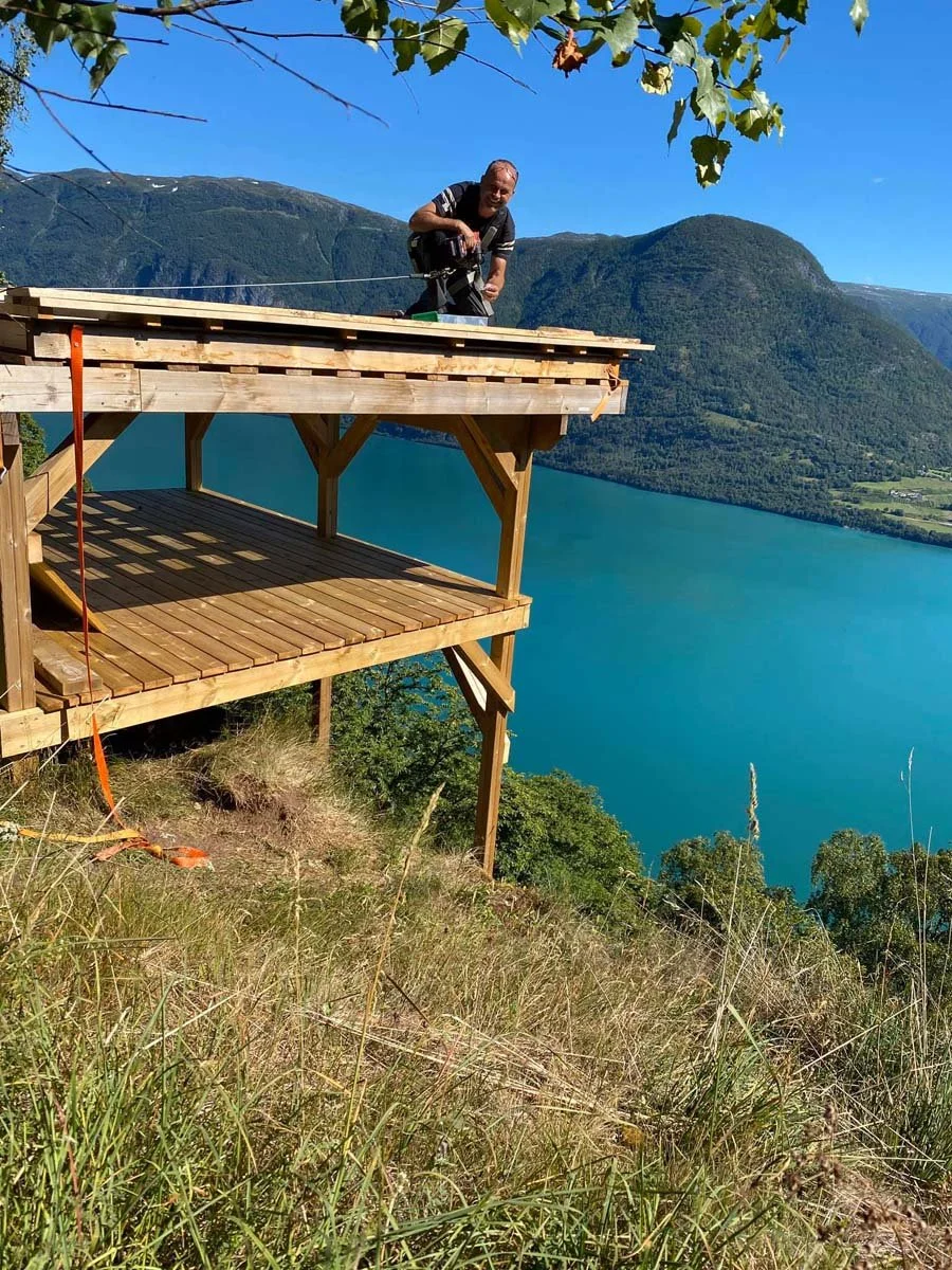 En mann oppå en treboden med utsikt over en innsjø omgitt av fjell.