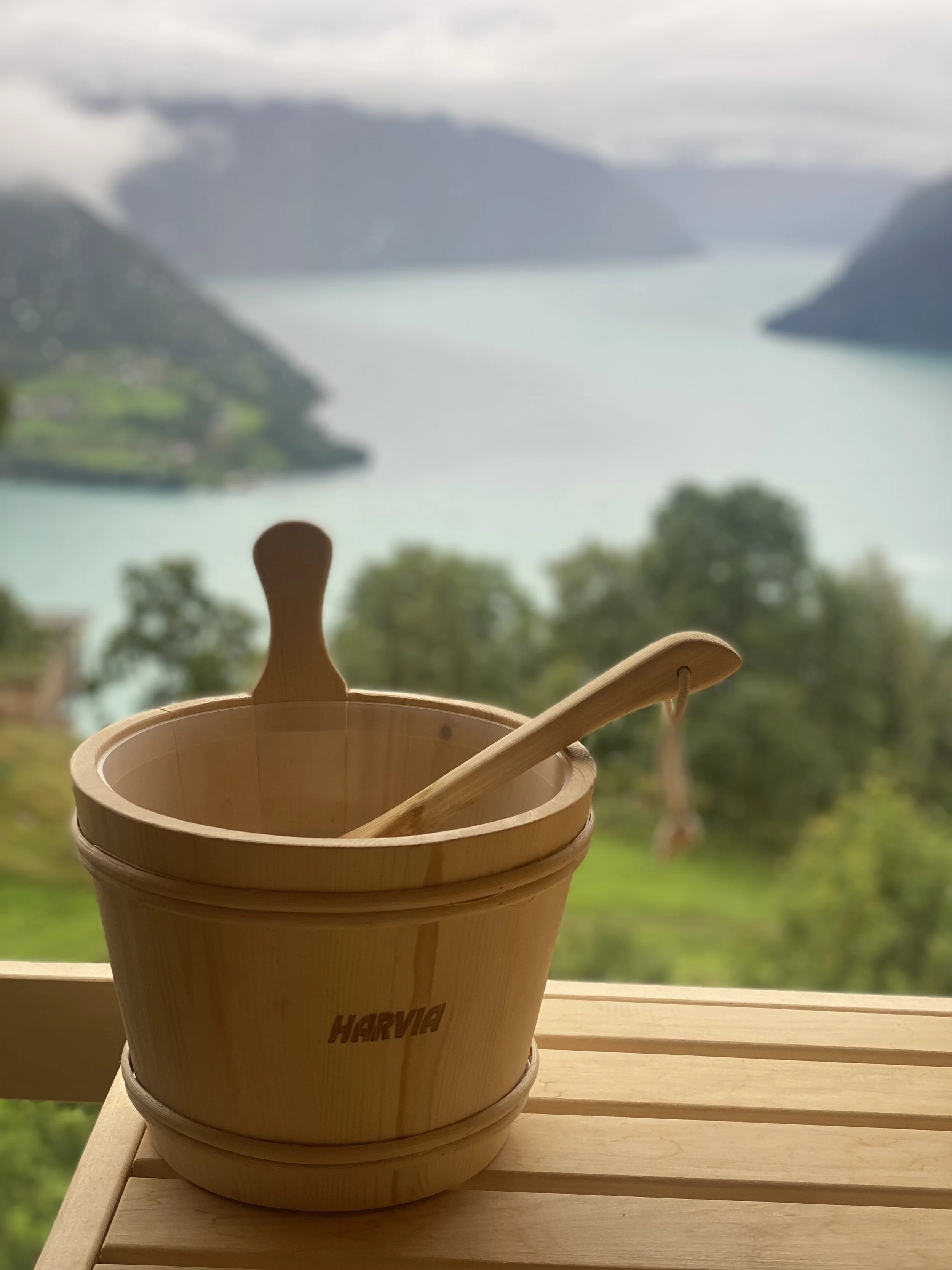 En vedfyrt badstuebøtte med lokk og treskaft, plassert på et trebord, med en landskap av innsjø og fjell i bakgrunnen.