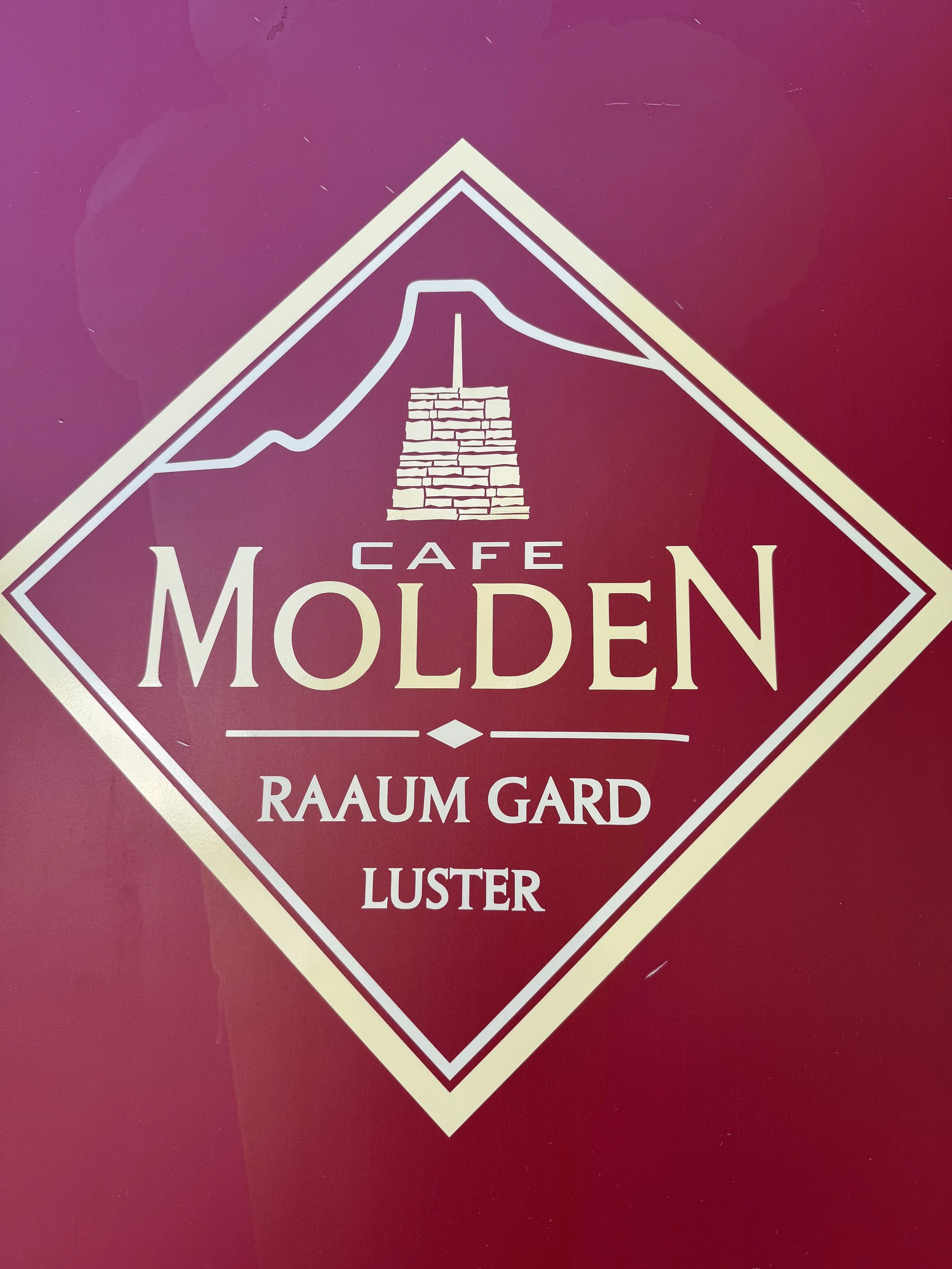 Et logo for Café Molden som viser en fjelltopp med en varde, med teksten «Café Molden», «Raum Gård», og «Luster».