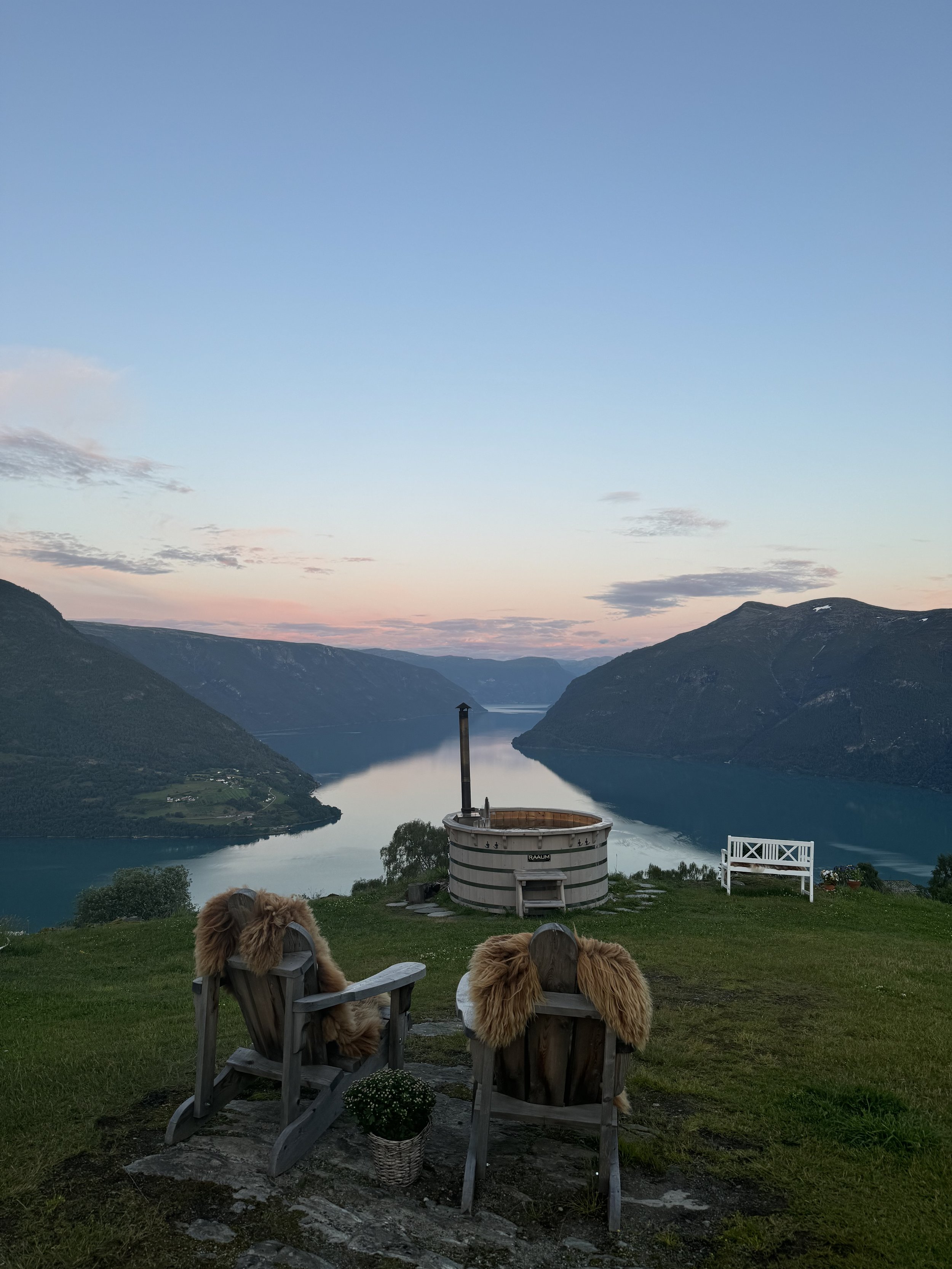 To tre stoler med pelsdeksler foran en innsjø omringet av fjell, med en badestamp i midten og en benk til høyre, under en rosa og blå himmel ved solnedgang.