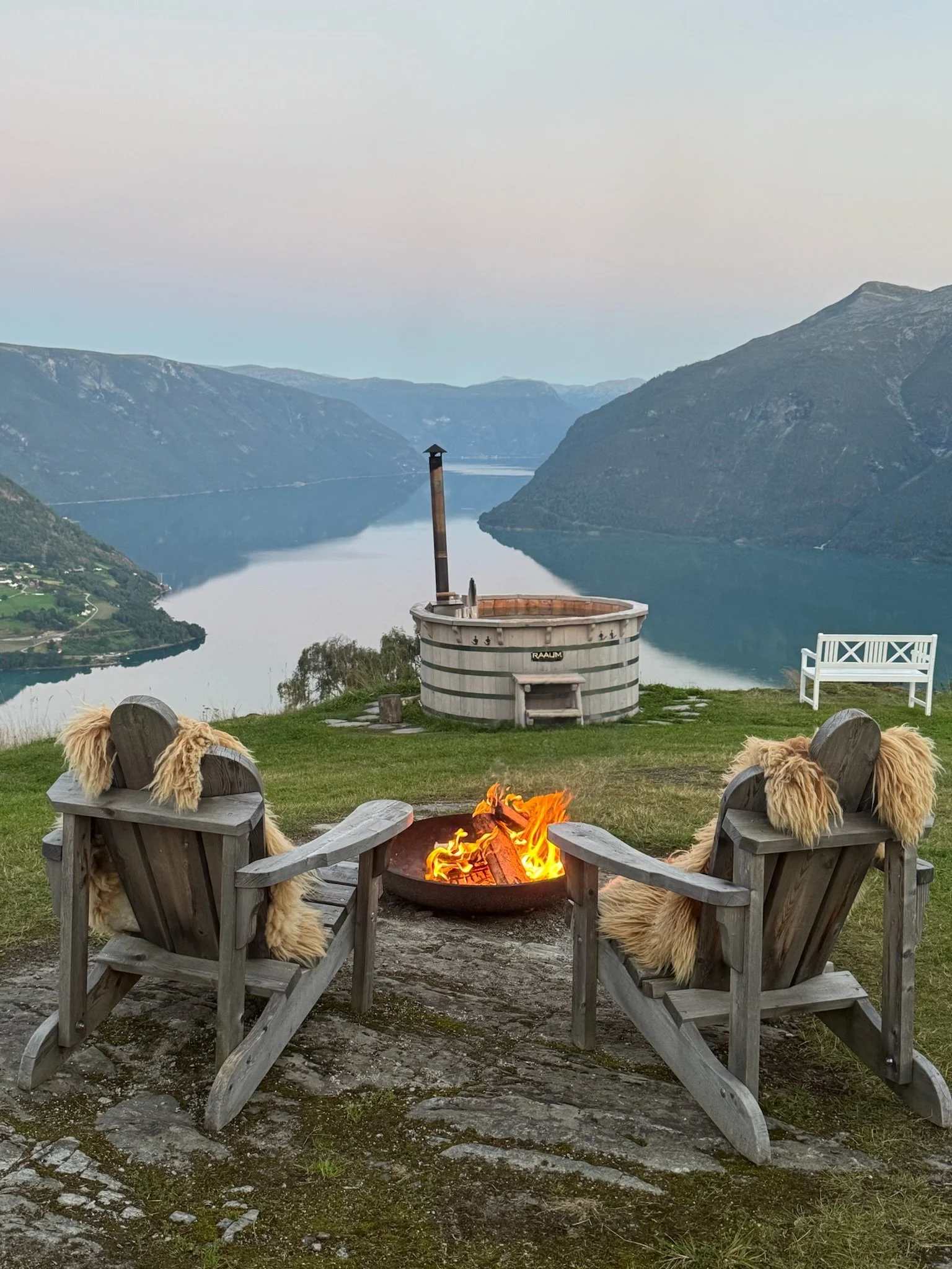 Hytteområde med fjord utsikt, to trestoler med saueskinn, et bål, badestamp og en benk