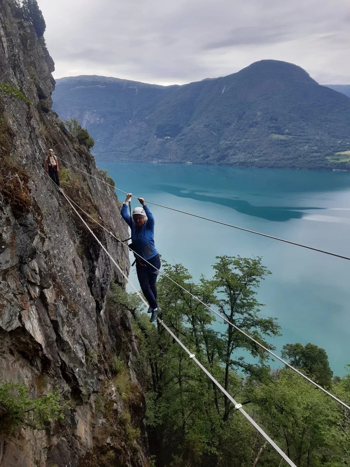 To personer går på en fjellkabel over en innsjø omgitt av fjell og skog, én person er nærmest kamera og holder fast i kabelen, den andre følger bak.