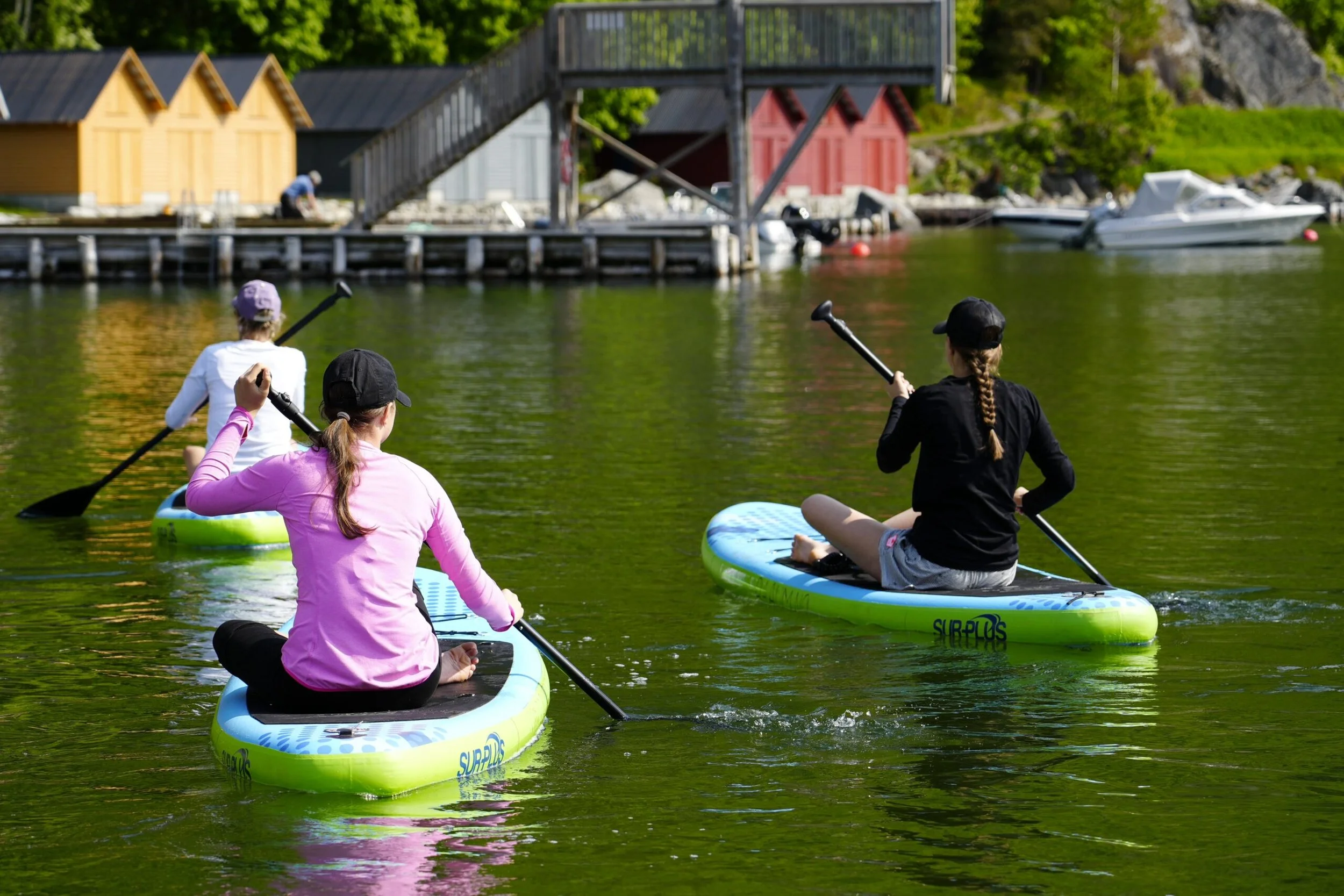 Tre personer som padler på vannet på SUP-brett, to kvinner og en jente, med små fargede hus og båter i bakgrunnen.