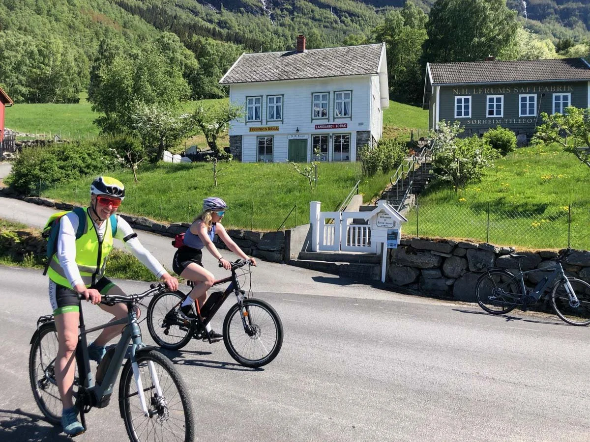To personer sykler på en vei med grønnsakser og hus i bakgrunnen. En av personene har hjelm og refleksvest.