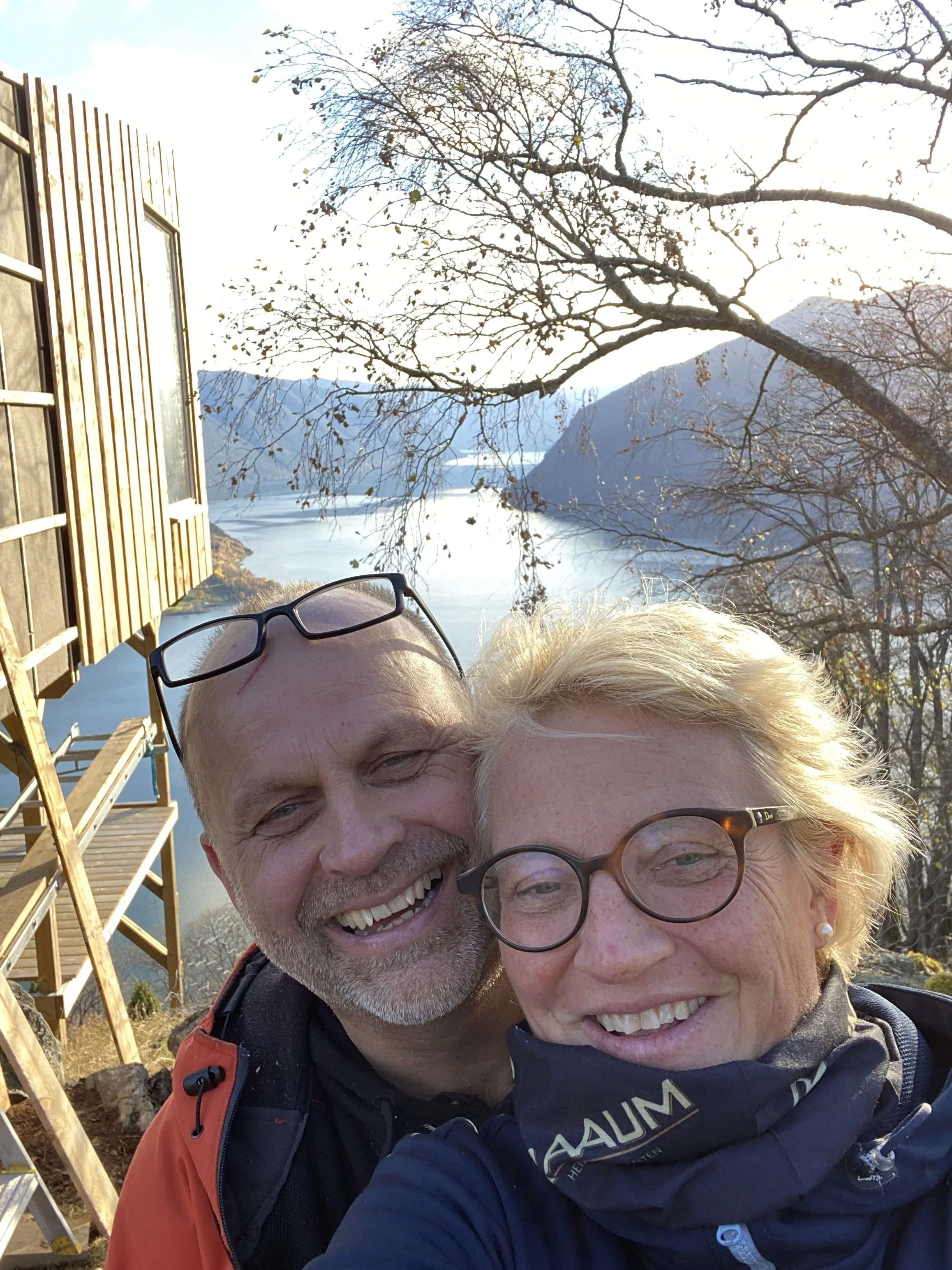 To glade personer tar en selfie på en utendørs sted med fjell, vann og trær uten blader i bakgrunnen, og et tre i forgrunnen.