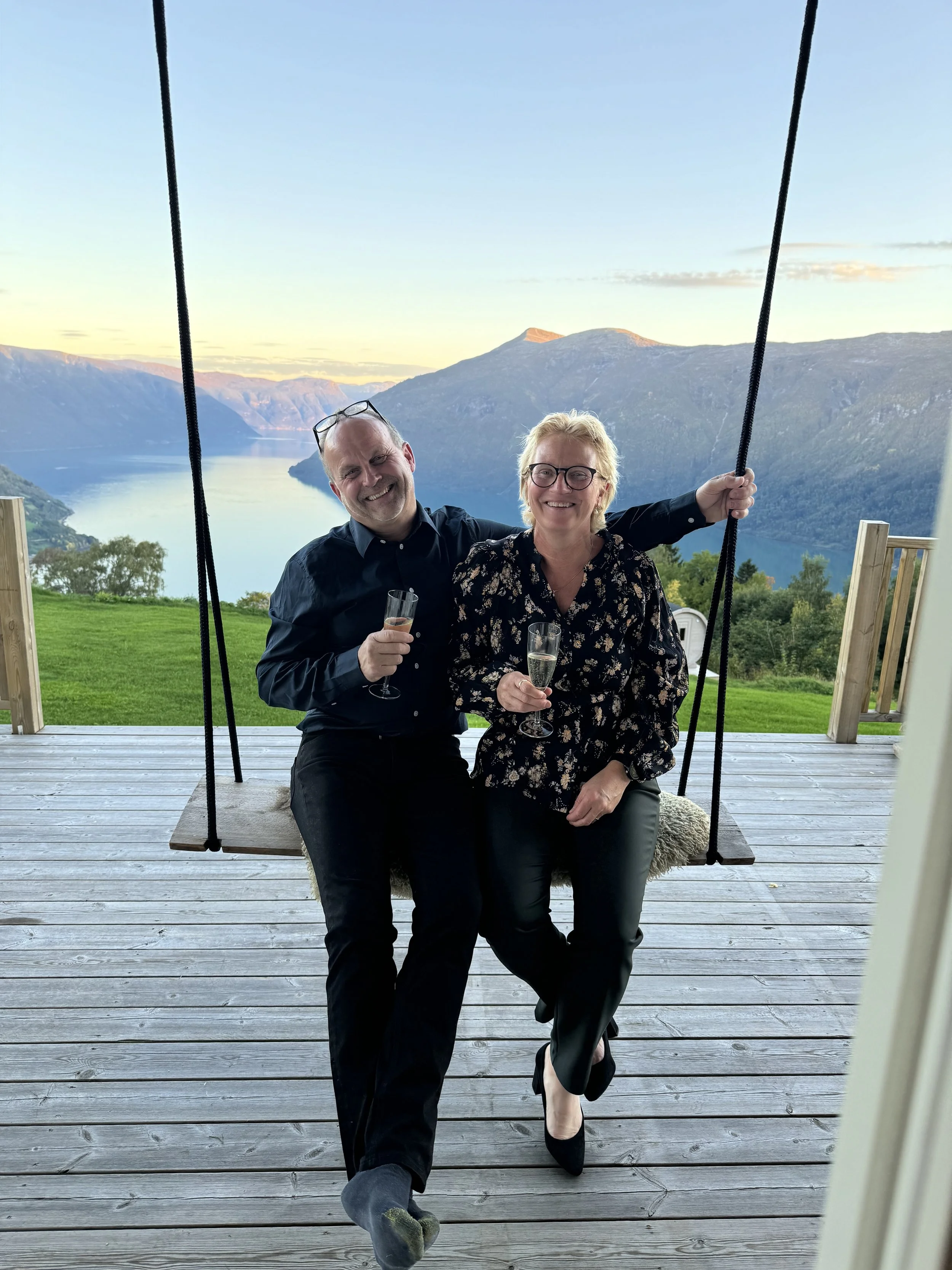 To personer, en mann og en kvinne, sitter på en hengende benk på en terrasse med utsikt over fjorder og fjell ved solnedgang, og holder champagneglass.
