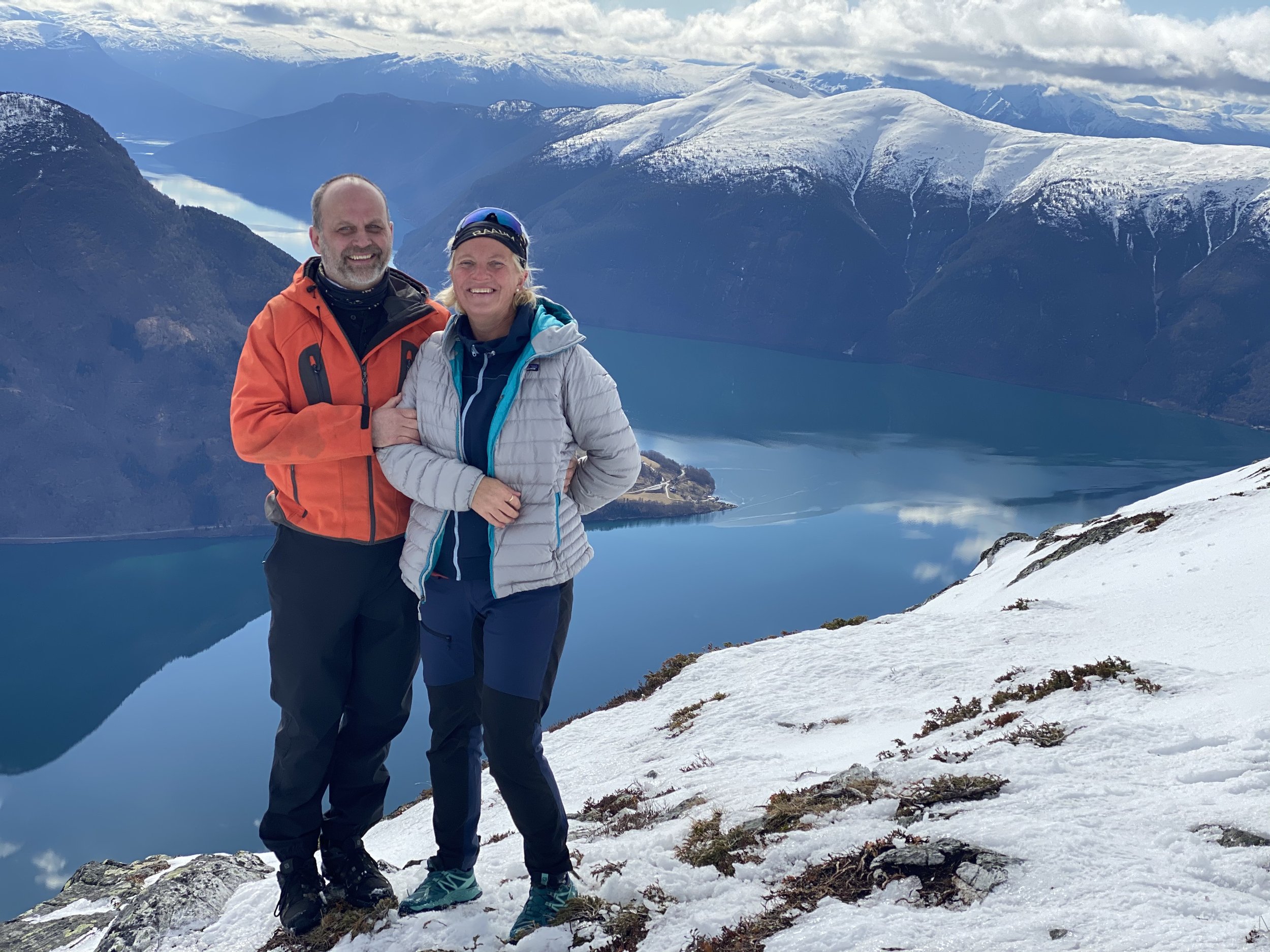 To personer på fjelltur, en mann i oransj jakke og en kvinne i grå jakke, står på snø med fjell og innsjø i bakgrunnen.