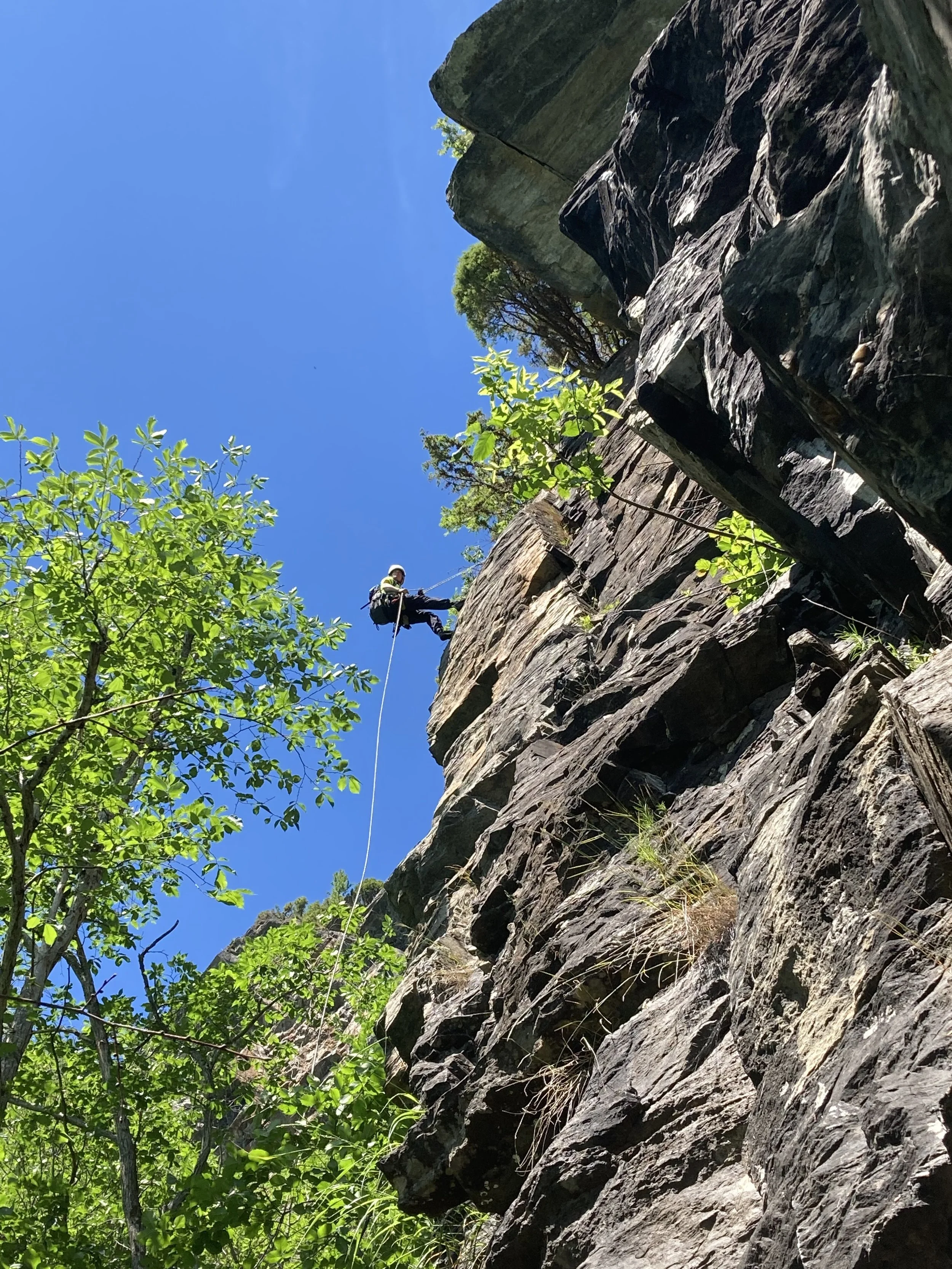 En person klatrer opp en fjellvegg med klatreutstyr under en klar blå himmel.
