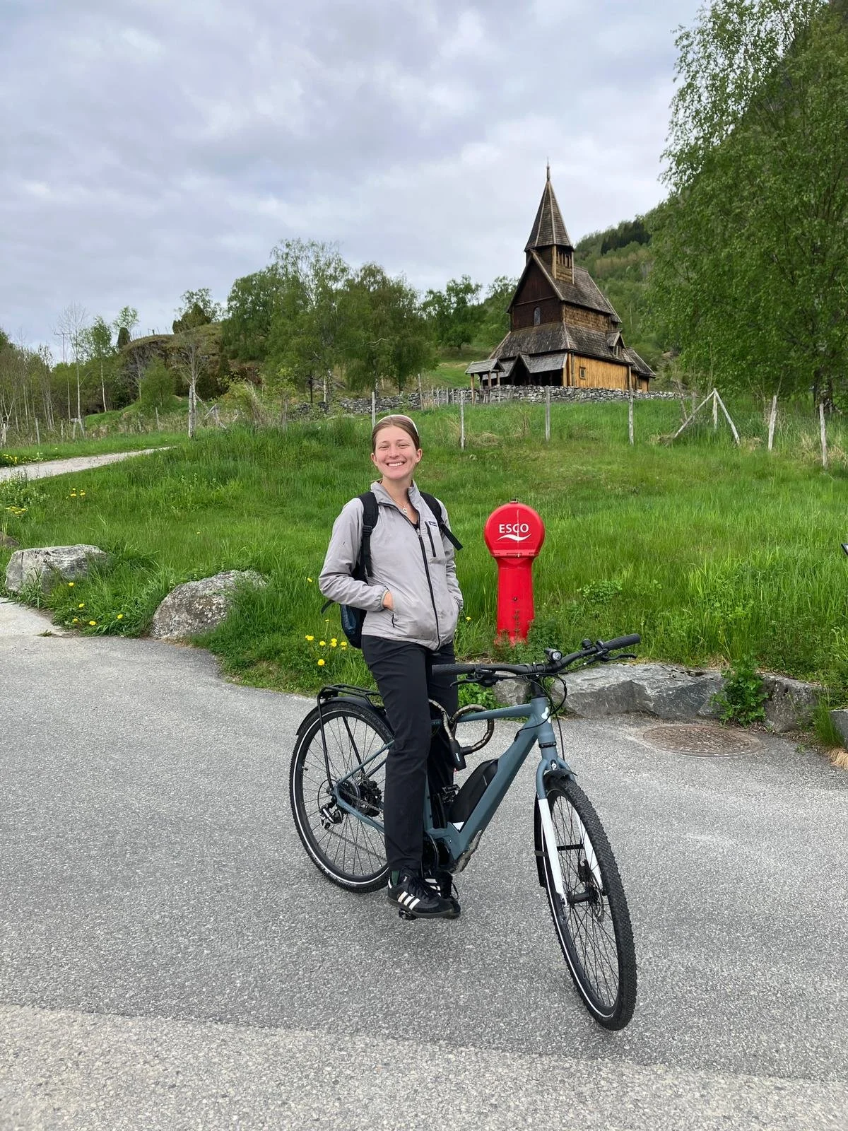 En kvinne med sykkel står på en vei med grønt landskap og en trebygning i bakgrunnen, under en overskyet himmel.