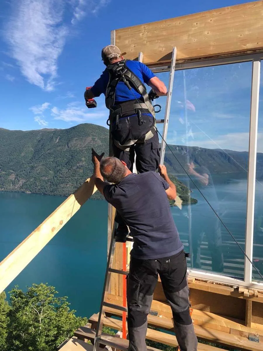 To håndverkere bygger et hus utendørs med utsikt over vann og fjell, en av dem står på en vei av tre med en trapp og på- og foran et glassvindu, en av dem holder en skrutrekker.
