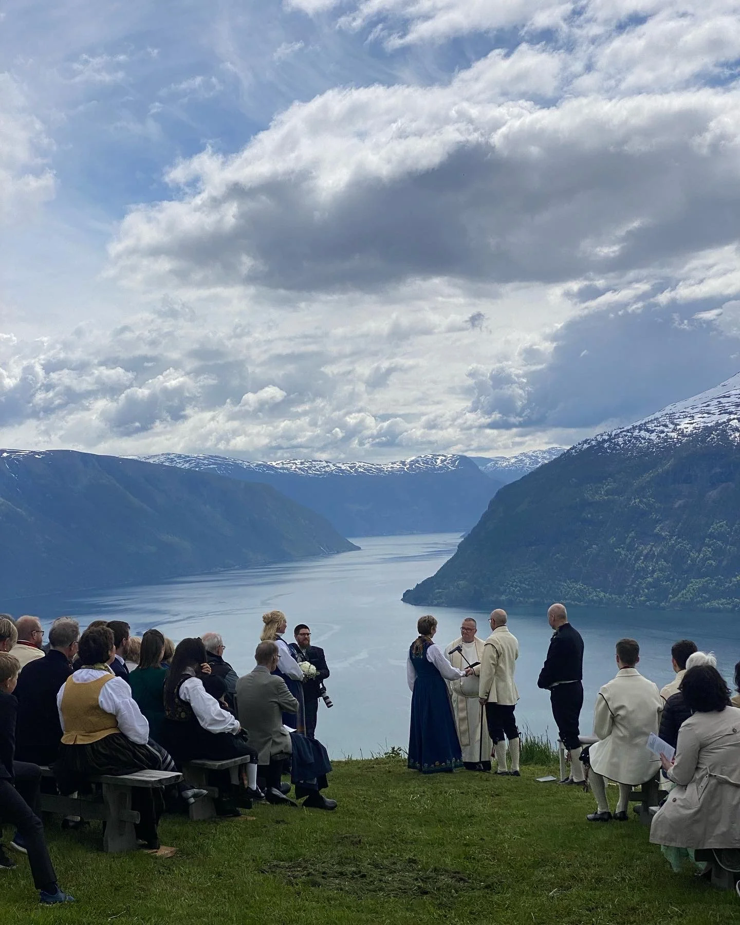 Bryllupseremoni utendørs med fjell og fjord i bakgrunnen, personer i tradisjonell klede og formell påkledning.