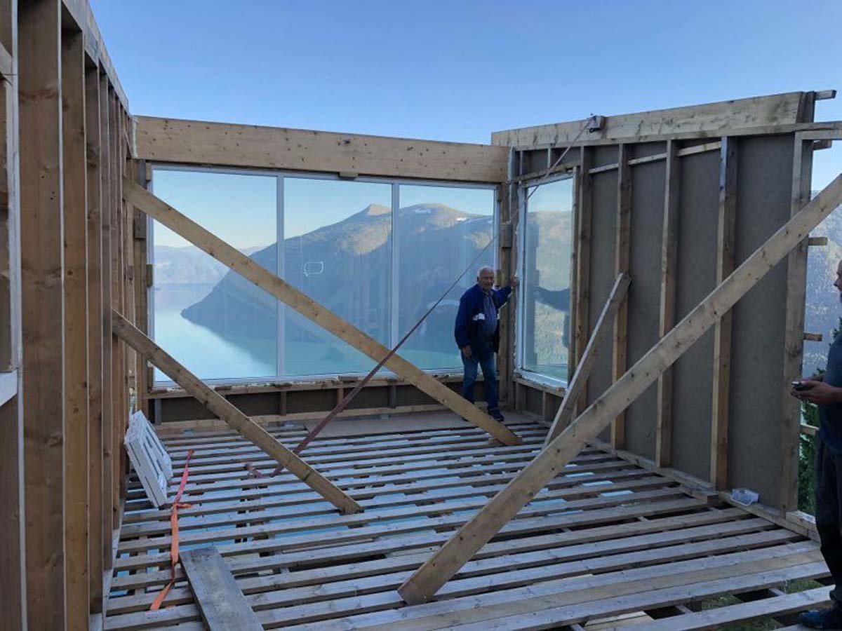 Byggearbeid innendørs med utsikt over fjell og innsjø, to personer som jobber, treverk og byggematerialer rundt.