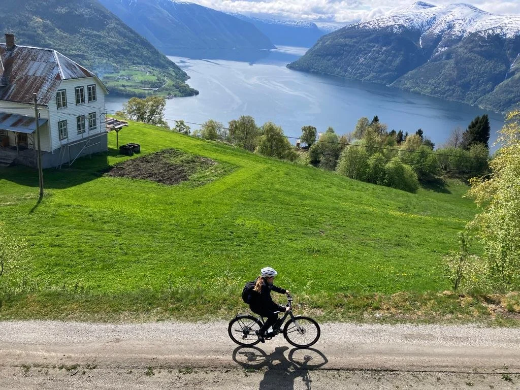 En person sykler på en grusvei ved en grønn ås med et hus, med fjord og fjell i bakgrunnen.