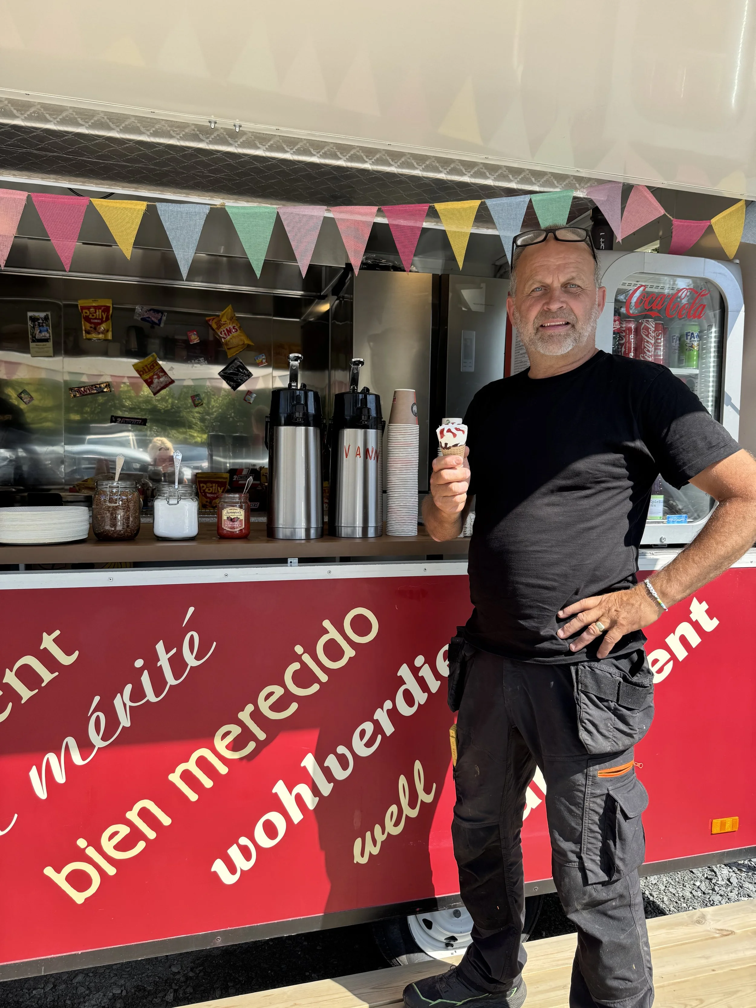 En mann som står foran en isvogn med kondimentflasker og kjeks, og holder is med sjokoladesirup. Mannen er iført svart t-skjorte og cargo bukser, og står på en treplate.