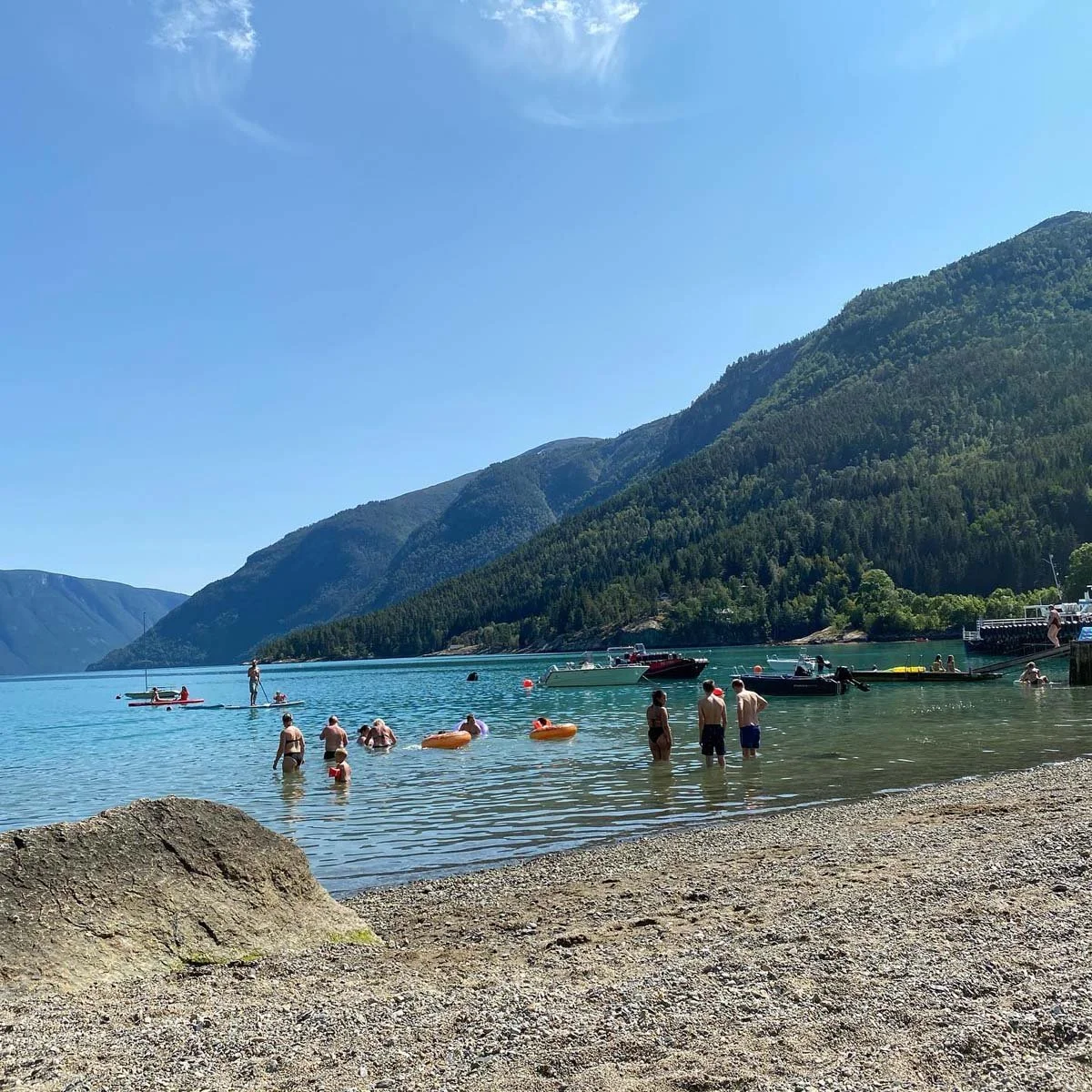 Folk svømmer og leker i en innsjø omgitt av fjell, med båter og mennesker på stranden.