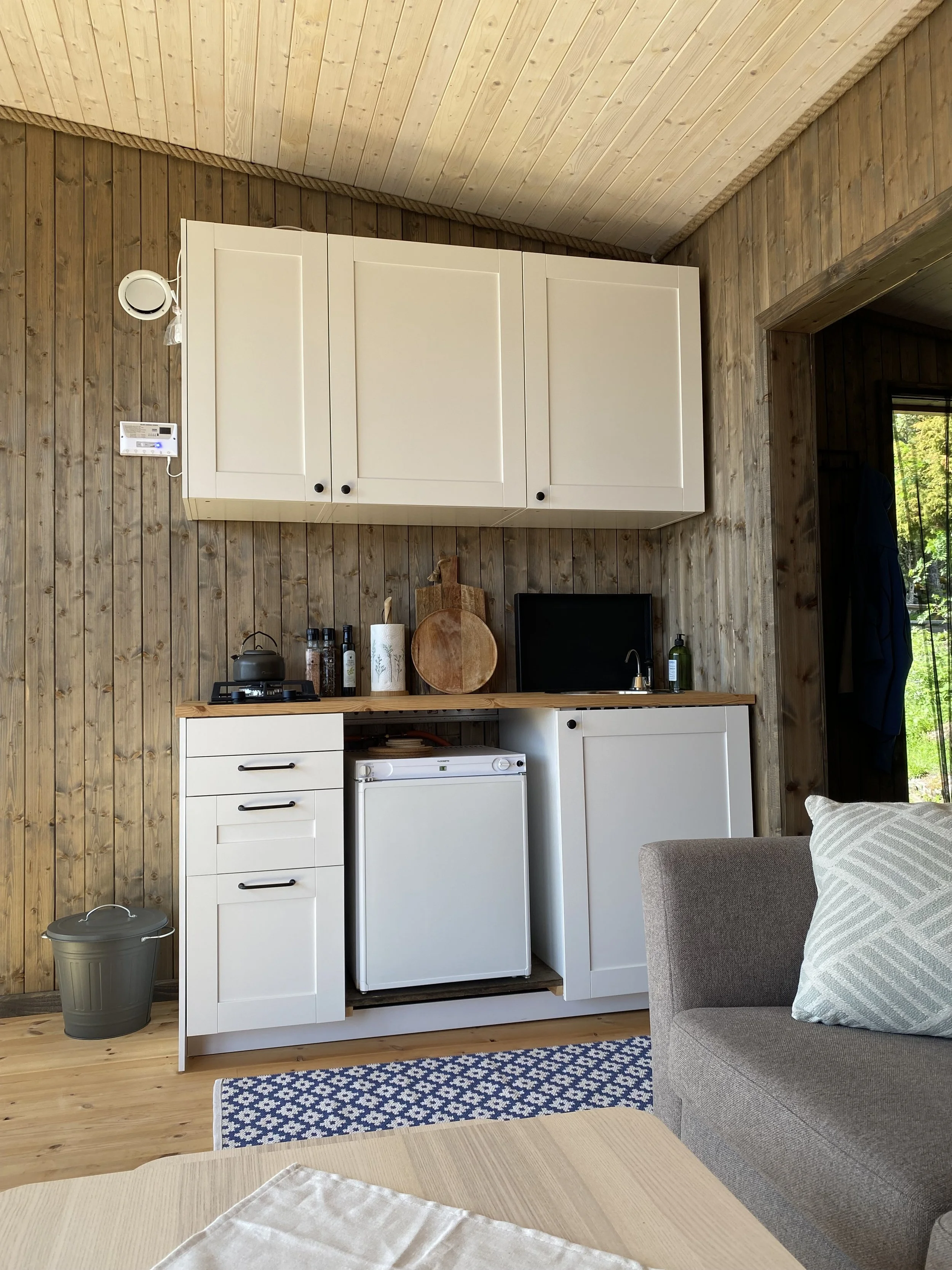 Kjøkkenområde med hvite skap, benkeplate i tre, liten vask, en mini fryser, og ulike kjøkkenutstyr, mot en vegg av trepanel.