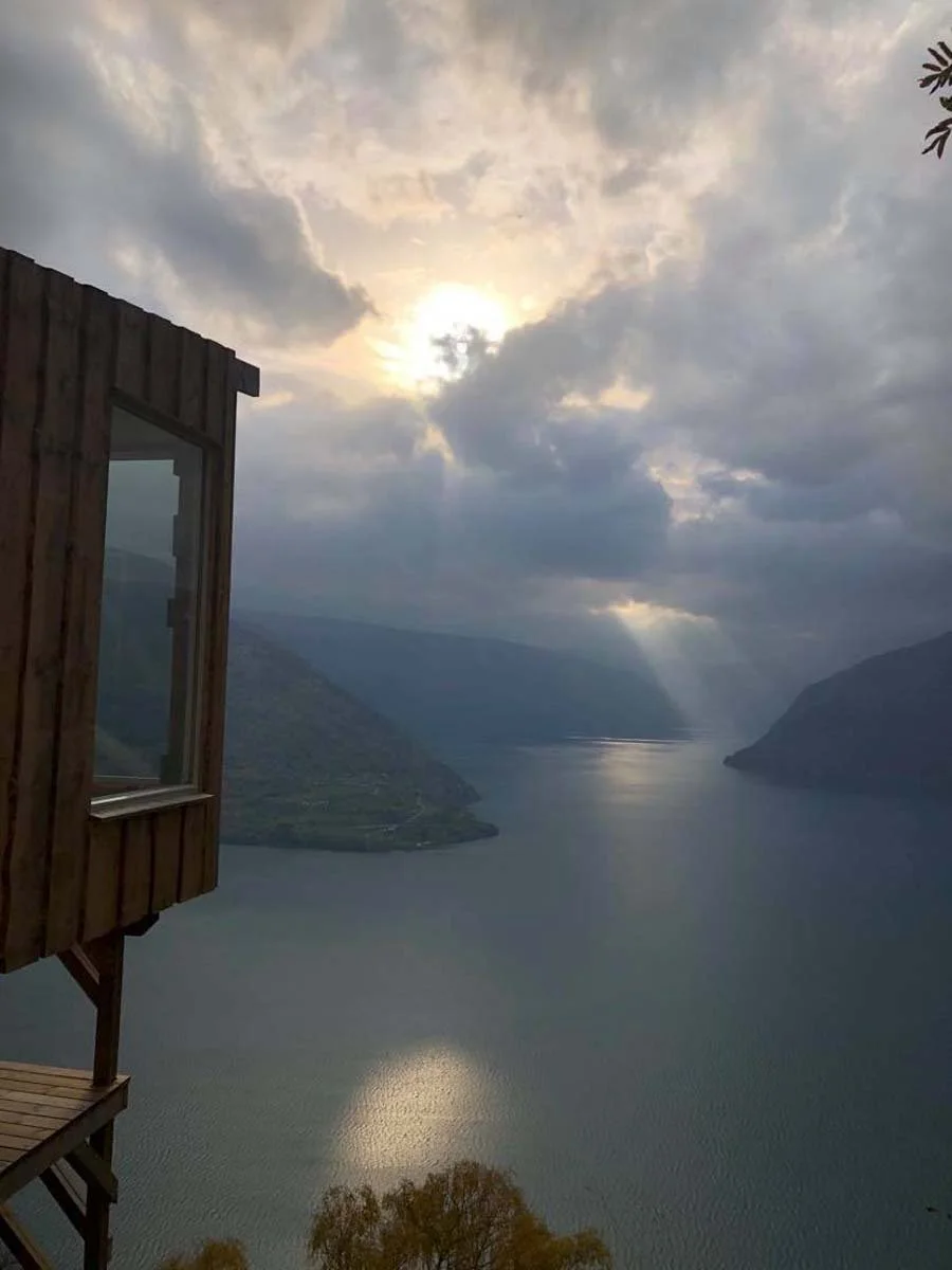 Utsikt over en innsjø omgitt av fjell, sett fra en trestue eller hytte, med et åpent vindu i venstre hjørne og en himmel med sol og skyer.