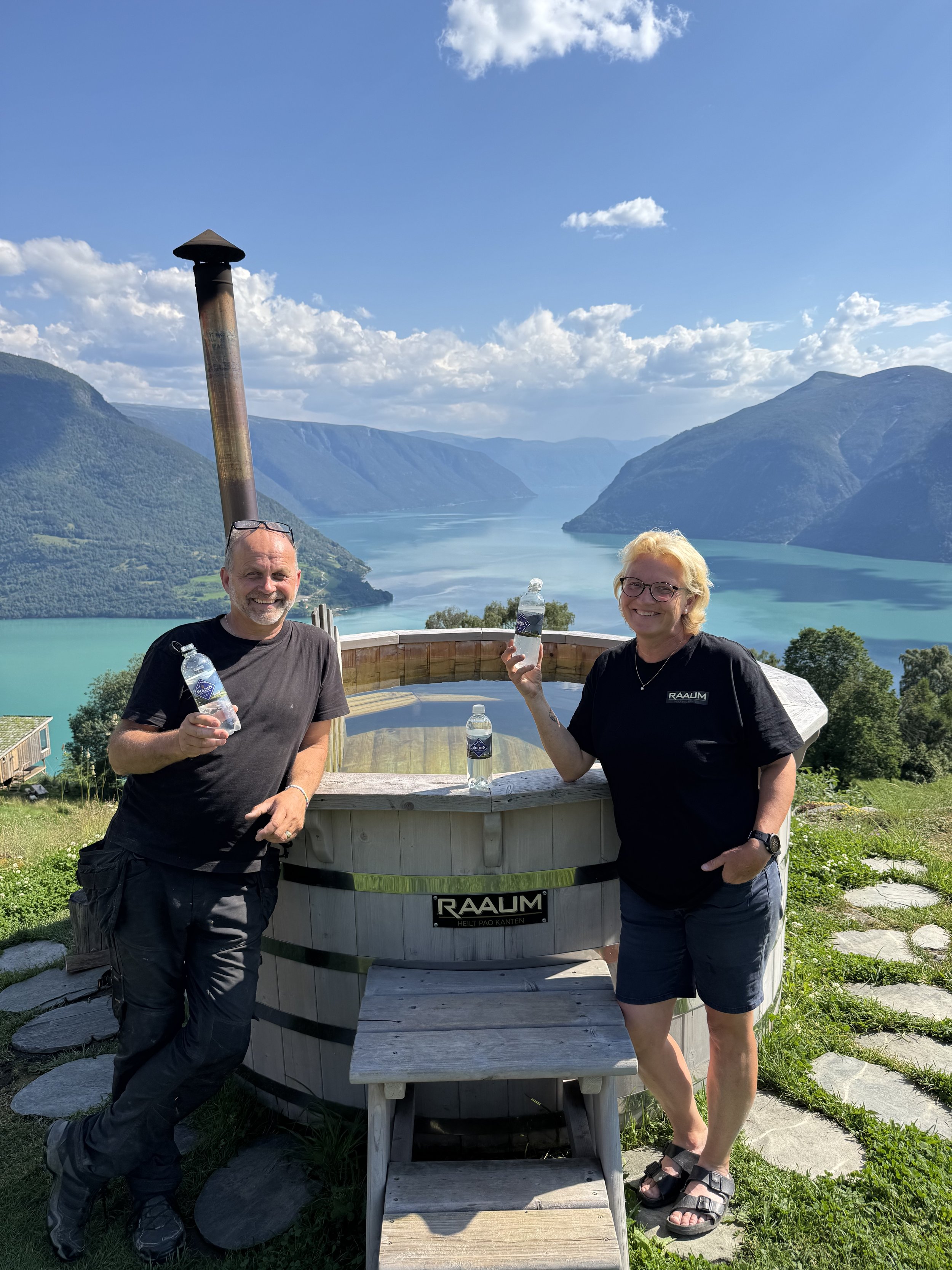 To personer med vannflasker foran en utendørs badestamp med fjell og innsjø i bakgrunnen.