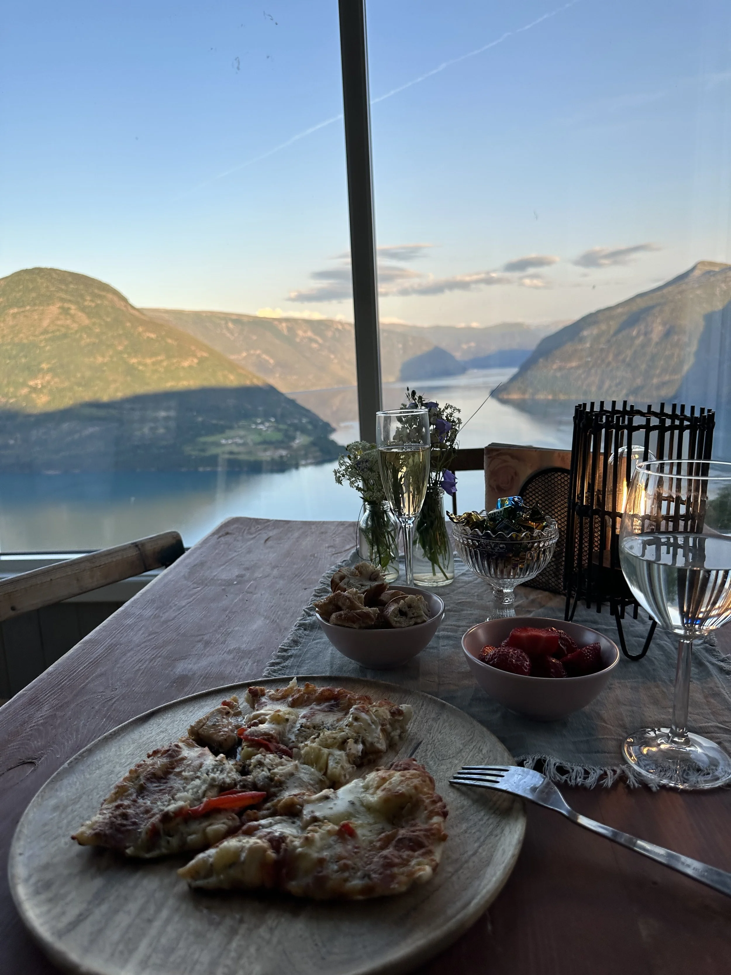 Middagsscenario med pizza på tre-fire stykker, en bolle med jordbær, en bolle med snacks, to glass med hvitvin, og et lite blomsterarrangement på et bord med fjell og fjord i bakgrunnen.