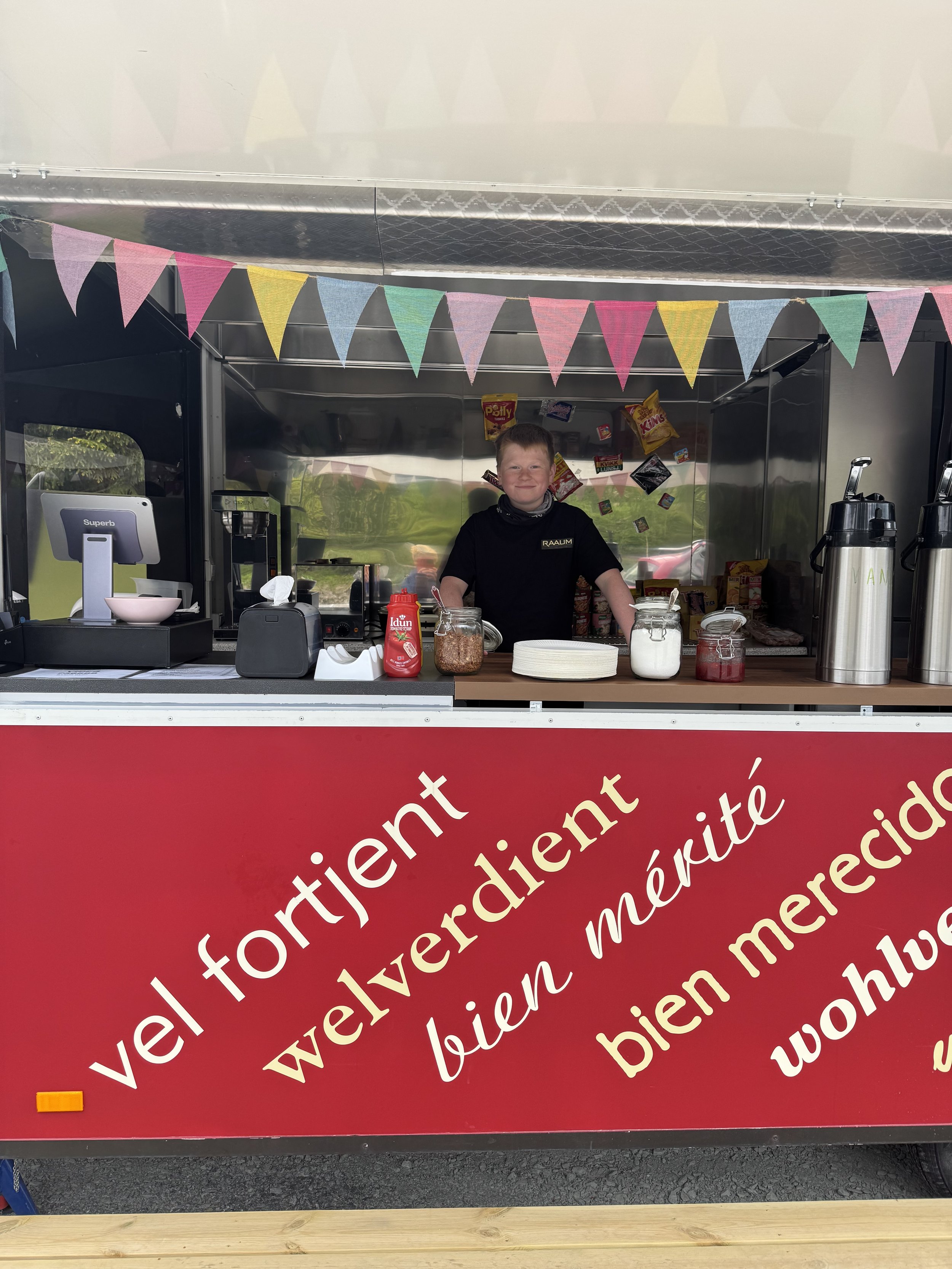 En liten gutt som står bak en salgsbod med fargerike flaggbånd hengende over. Bodsiden har tekst på norsk med ulike velkomsthilsener. Det er ulike matvarer og drikkevarer på disken.