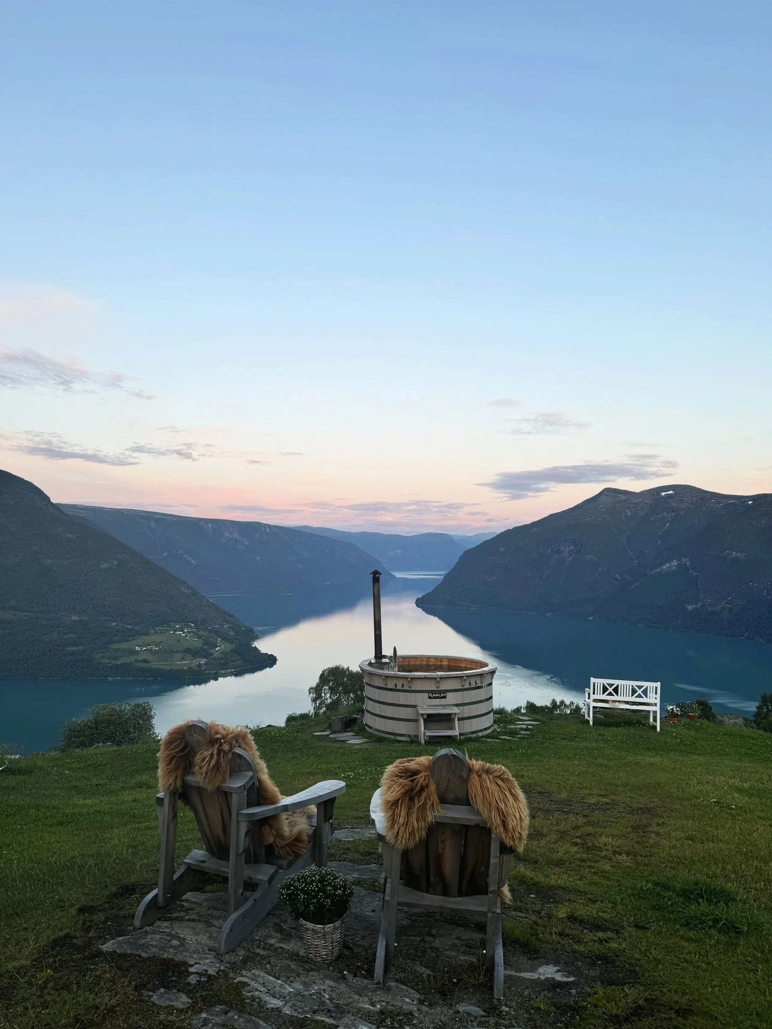To hunder i campingstoler med pelsdeksler foran en innsjø omgitt av fjell, med en badestamp og utsikt over vann og fjell i solnedgang