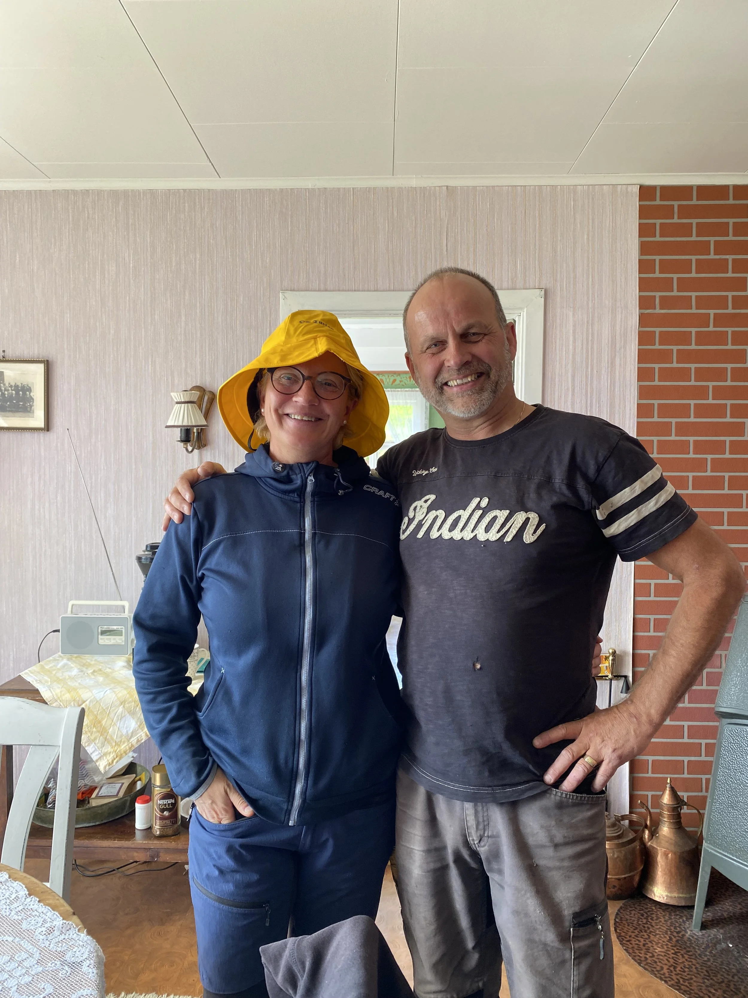 To personer smiler og står sammen innendørs, med en kvinne i blå jakke og gul hatt, og en mann i svart t-skjorte som sier «Indian».