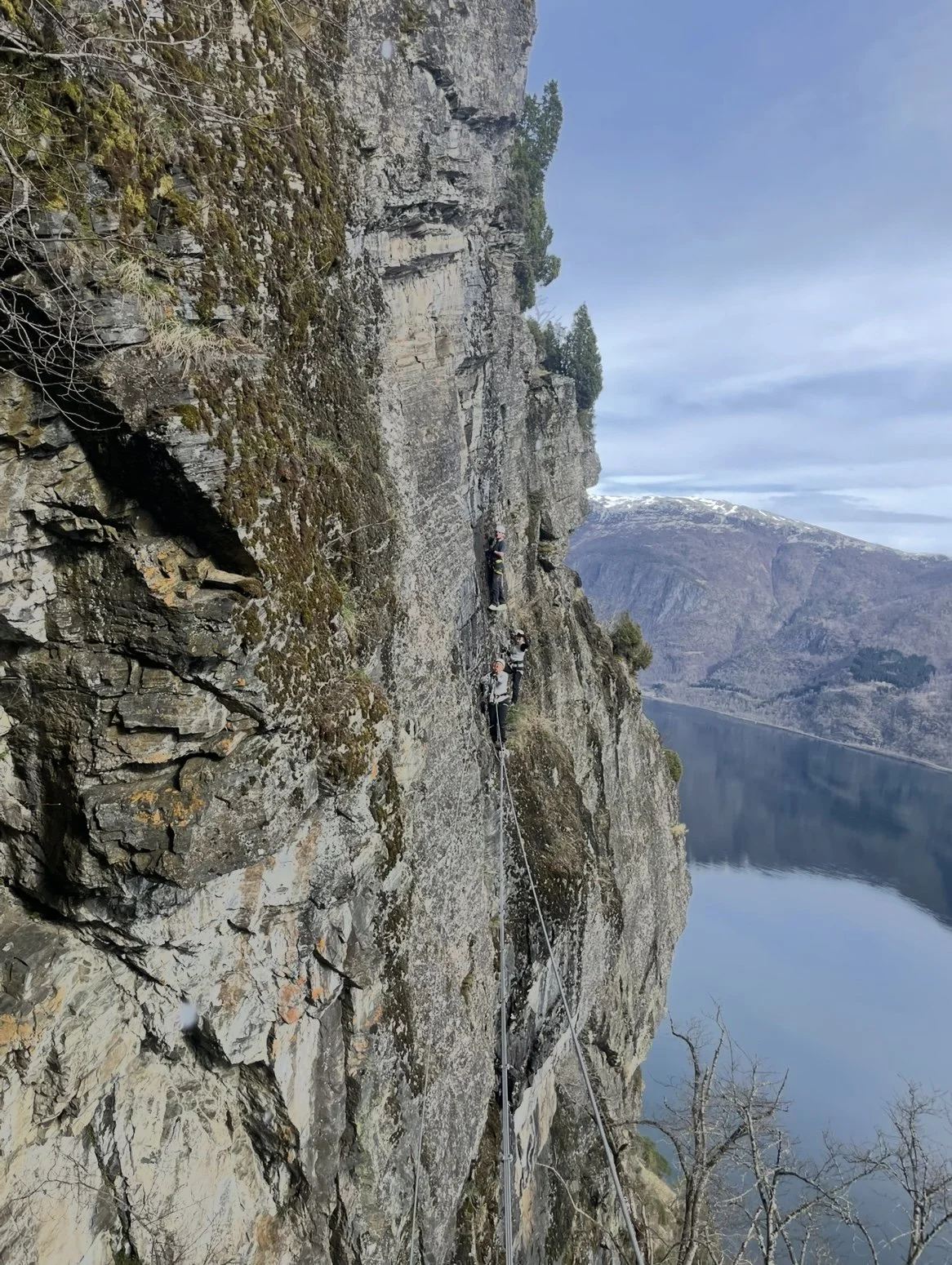 Fjellklatrere som klatrer på en vertikal fjellvegg med en vannkropp og fjell i bakgrunnen.