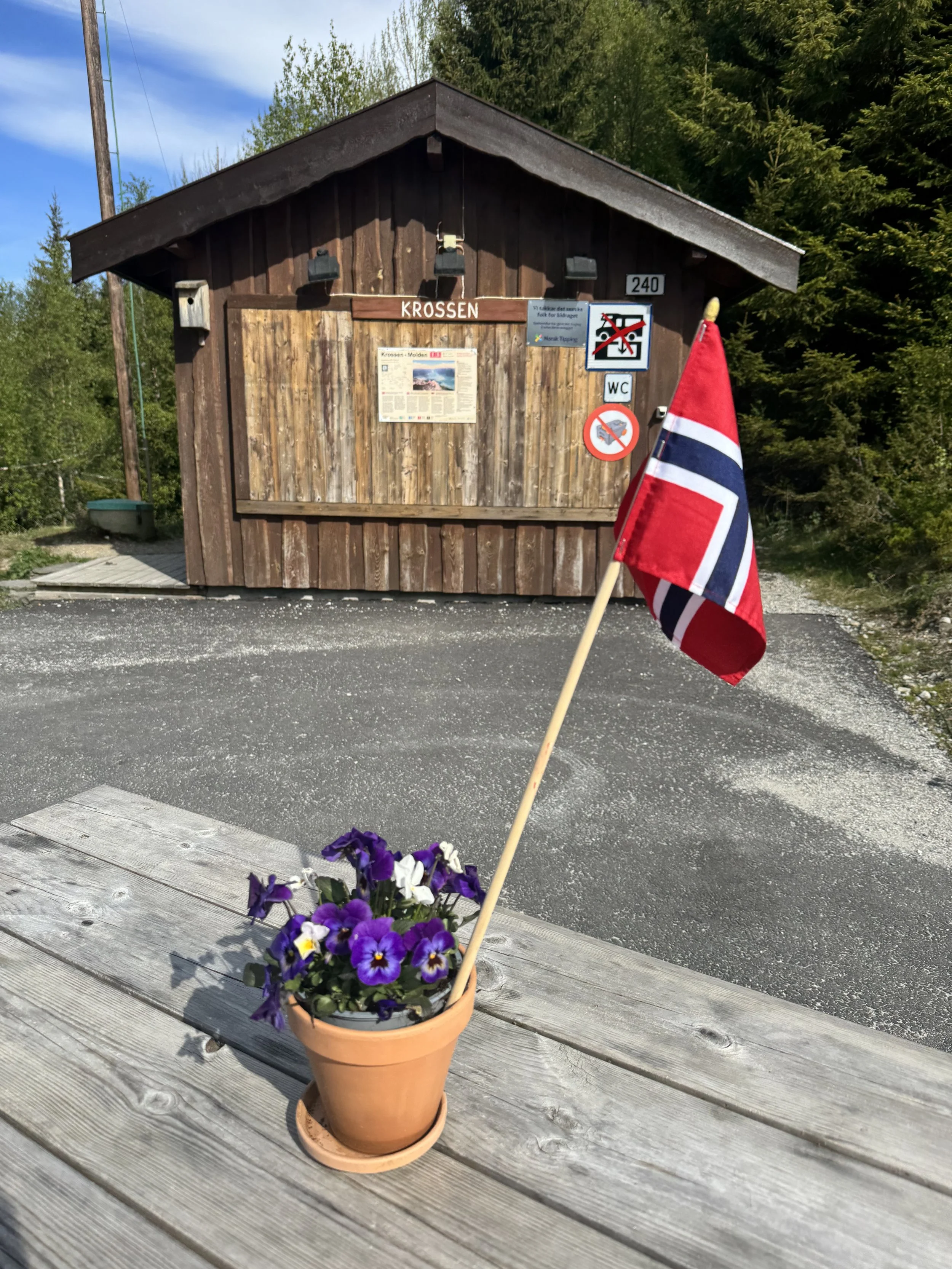 En liten hytte av tre med skilt som sier 'Krossen'. En norsk flagg i en blomsterpotte står foran hytta. Området er omgitt av grønne trær og en grusvei.