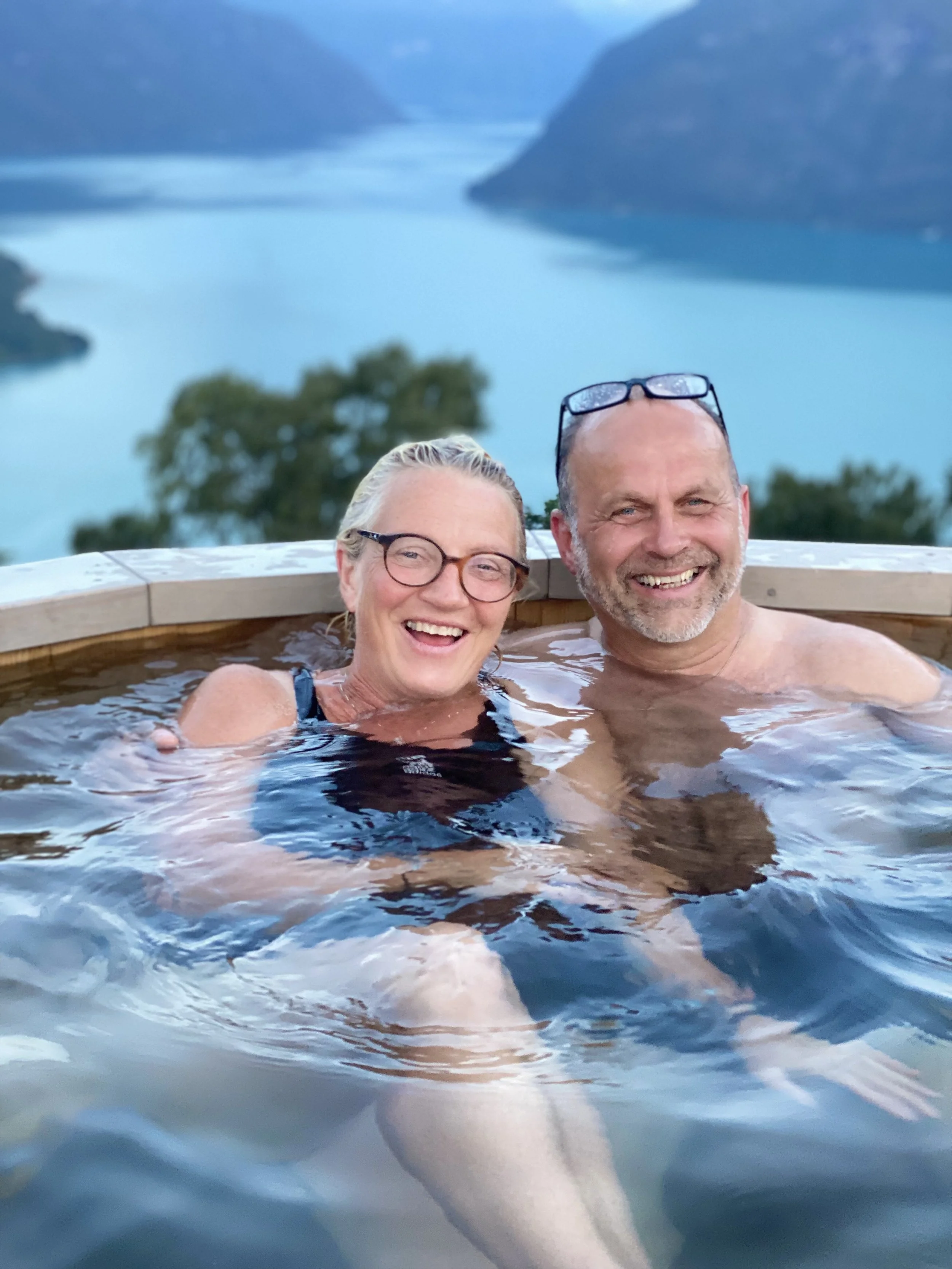 Et lykte av to voksne mennesker som smiler og koser seg i et boblebad med en innsjø eller fjellandskap i bakgrunnen.