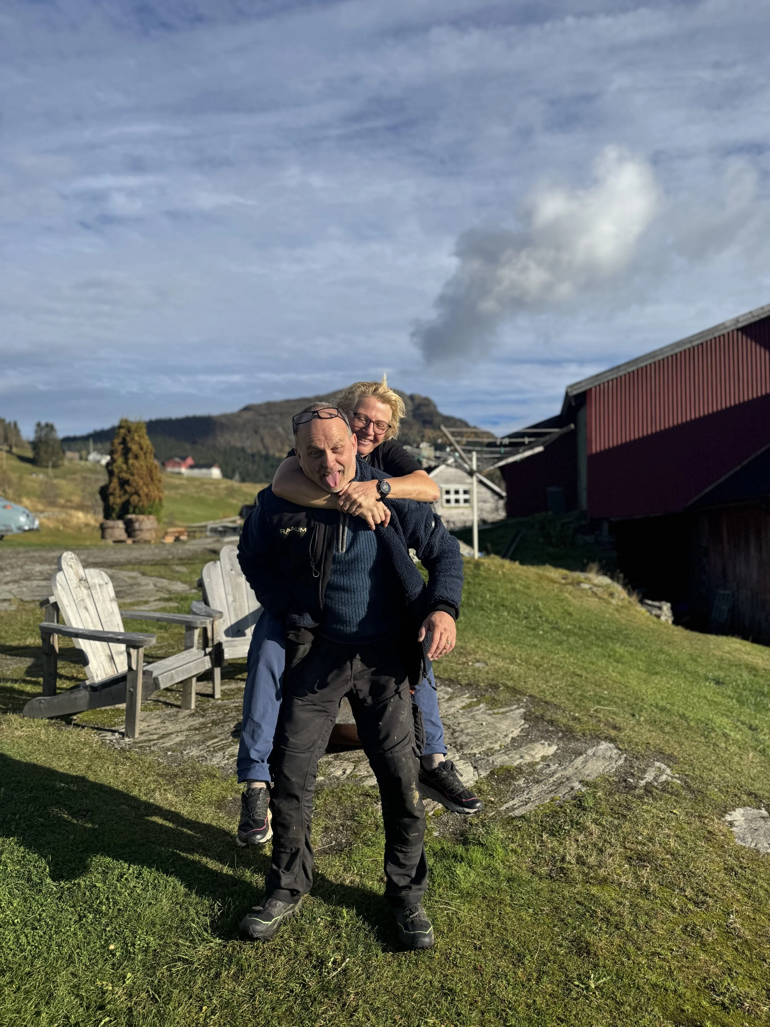 To personer som smiler og klemmer hverandre utendørs i et landskap med grastak, trær og hus, under en klar himmel med noen skyer.