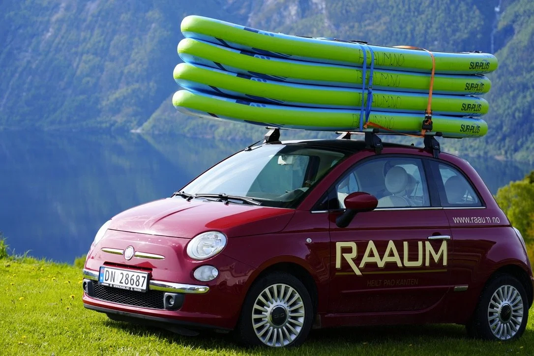 En rød Fiat 500 med surfebrett på taket, parkert på gress med fjellbakgrunn.