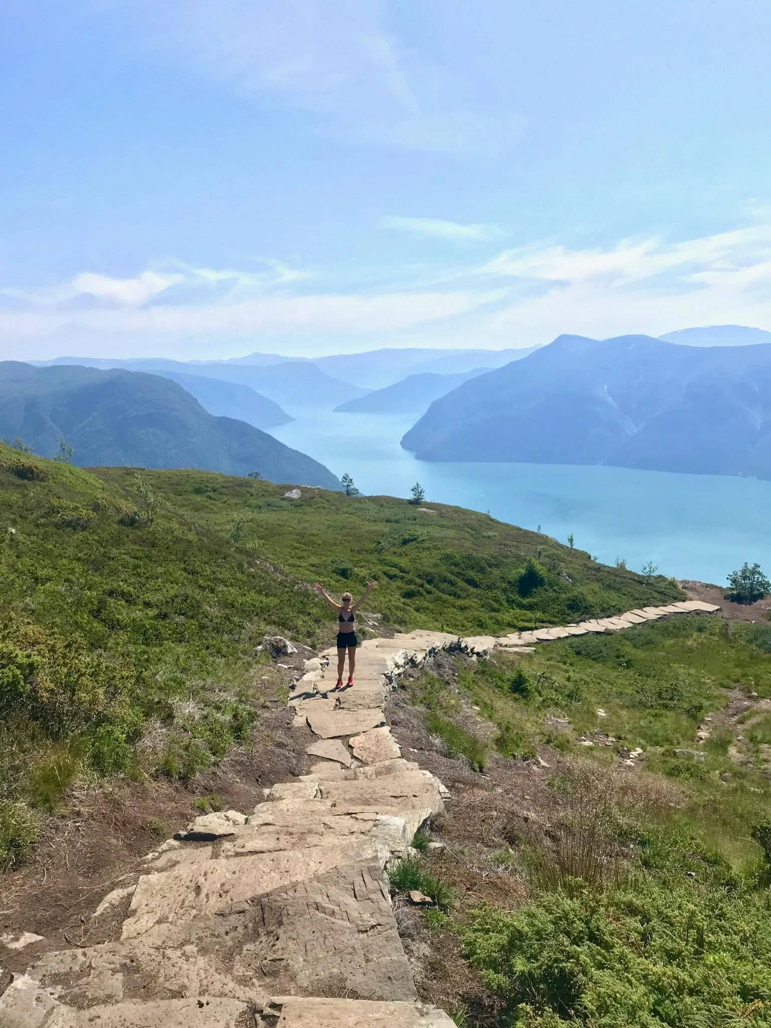 En person med armene hevet opp på en steinsti i fjellet med utsikt over en innsjø og fjell i bakgrunnen.