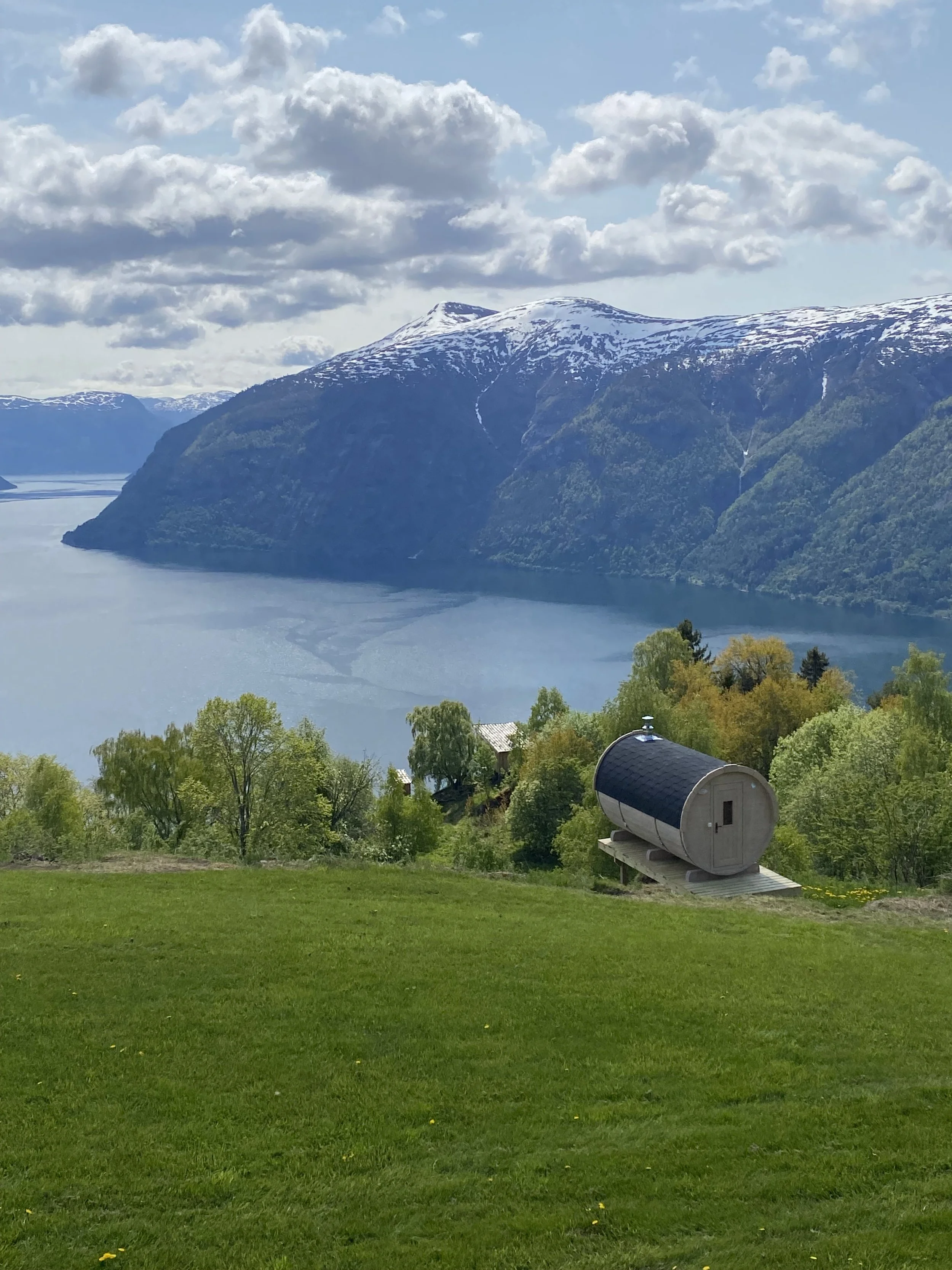 Grønt landskap med gress, en tønneformet badstue, trær og en fjord med fjell i bakgrunnen, snø på fjelltoppene, og en del av en himmel med skyer.