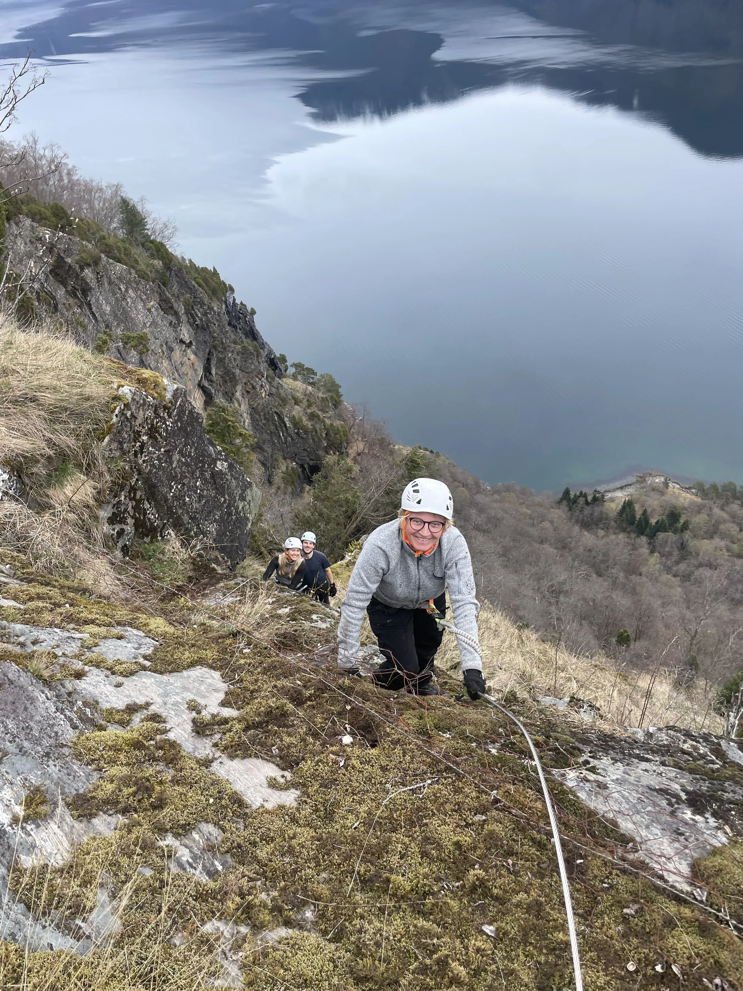Fire personer klatrer opp en bratt fjellside med hjelp av tau, med et stort vann og fjell i bakgrunnen.