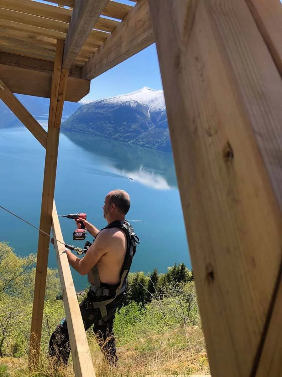 En mann med beskåret topp og utsikt over en fjord, fjell med snø, trerammer av tårn eller konstruksjon i forgrunnen.