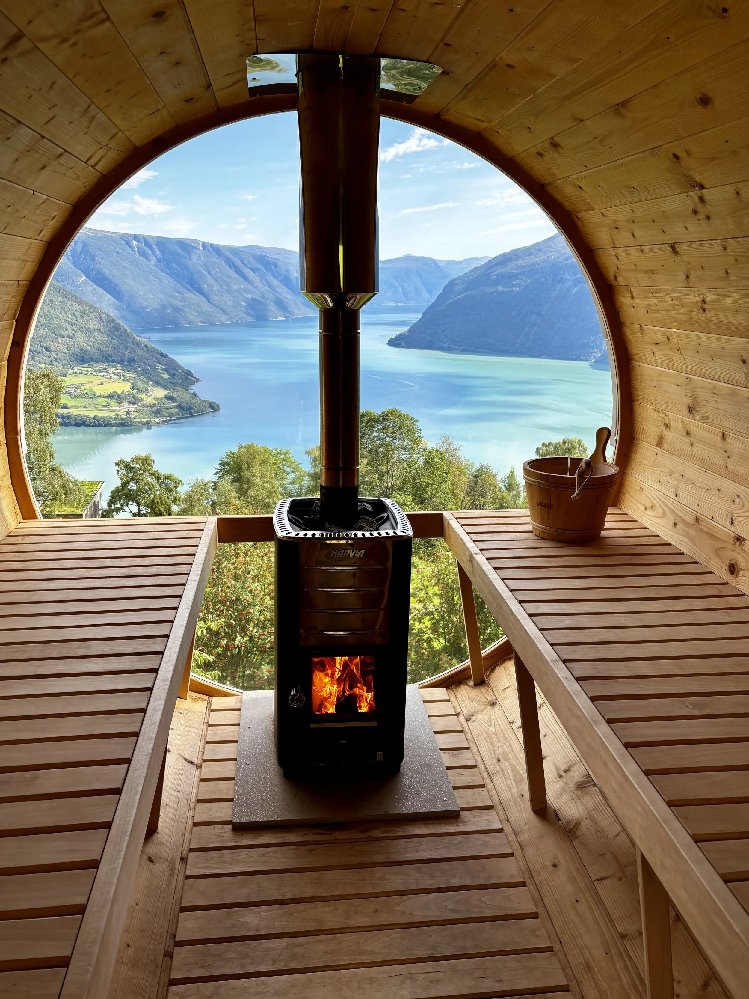 Utsikt fra en badstue med utsikt over en fjord og fjell, inne med trevegger, to trebenker, og en vedfyrt ovn.