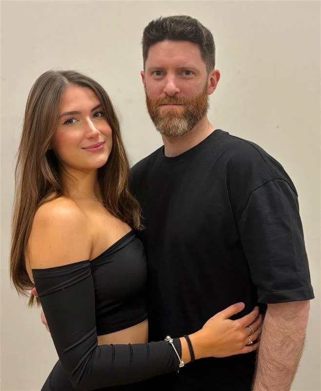 A young woman with long brown hair and a young man with short dark hair and a beard stand close together, smiling and looking at the camera against a plain background. The woman wears a black off-the-shoulder top, and the man wears a black t-shirt.