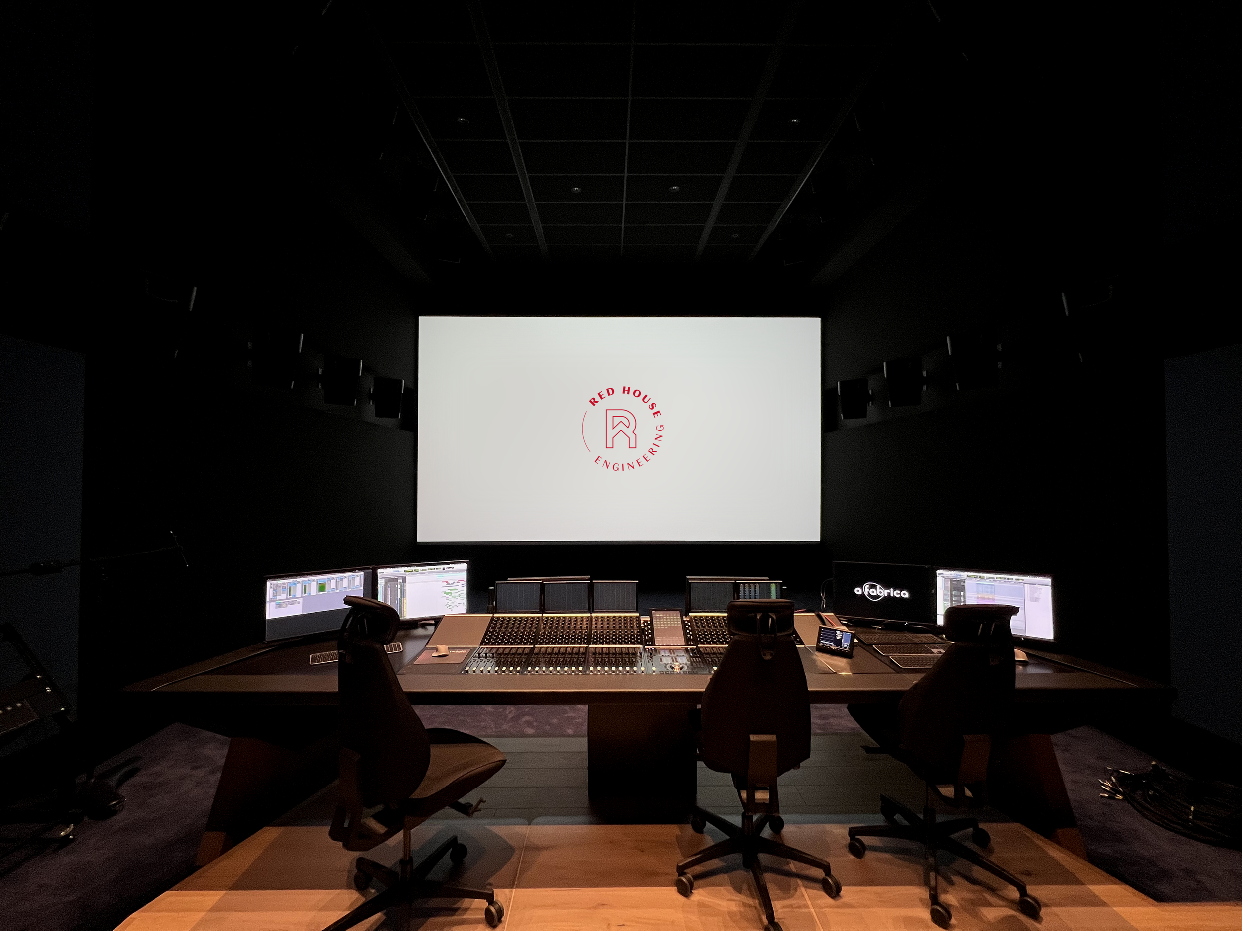 Salle de contrôle audio avec plusieurs écrans, un grand écran au fond affichant le logo 'Red House Engineering' et des chaises autour d'une console de mixage.