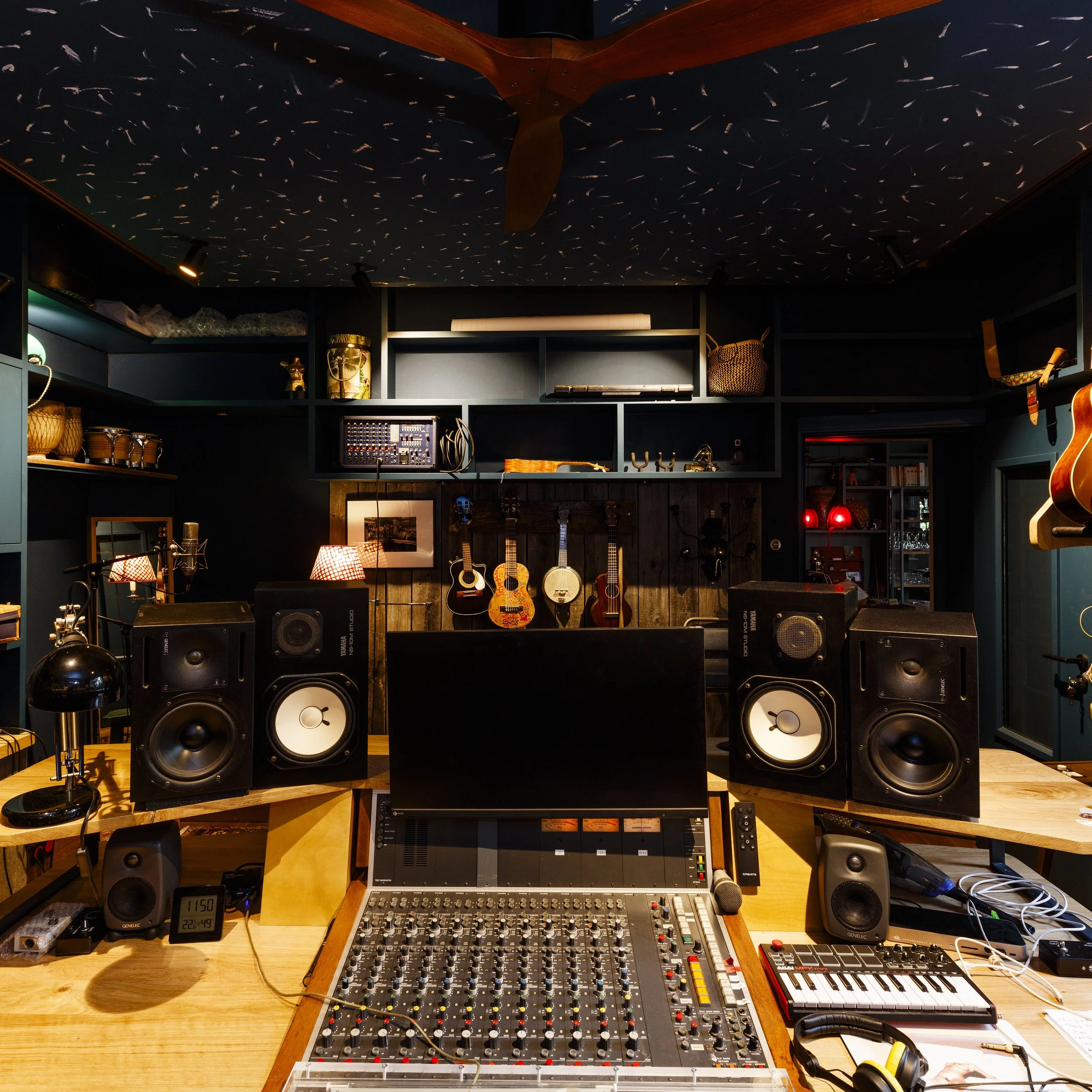 Une salle de studio d'enregistrement avec des instruments à cordes accrochés au mur, des enceintes, un ordinateur, un micro, un clavier MIDI, et des équipements audio. Des guitares sont suspendues au mur et des lampes produisent une lumière chaleureuse. Le plafond est décoré avec un ciel étoilé et un plafonnier en forme d'hélice.