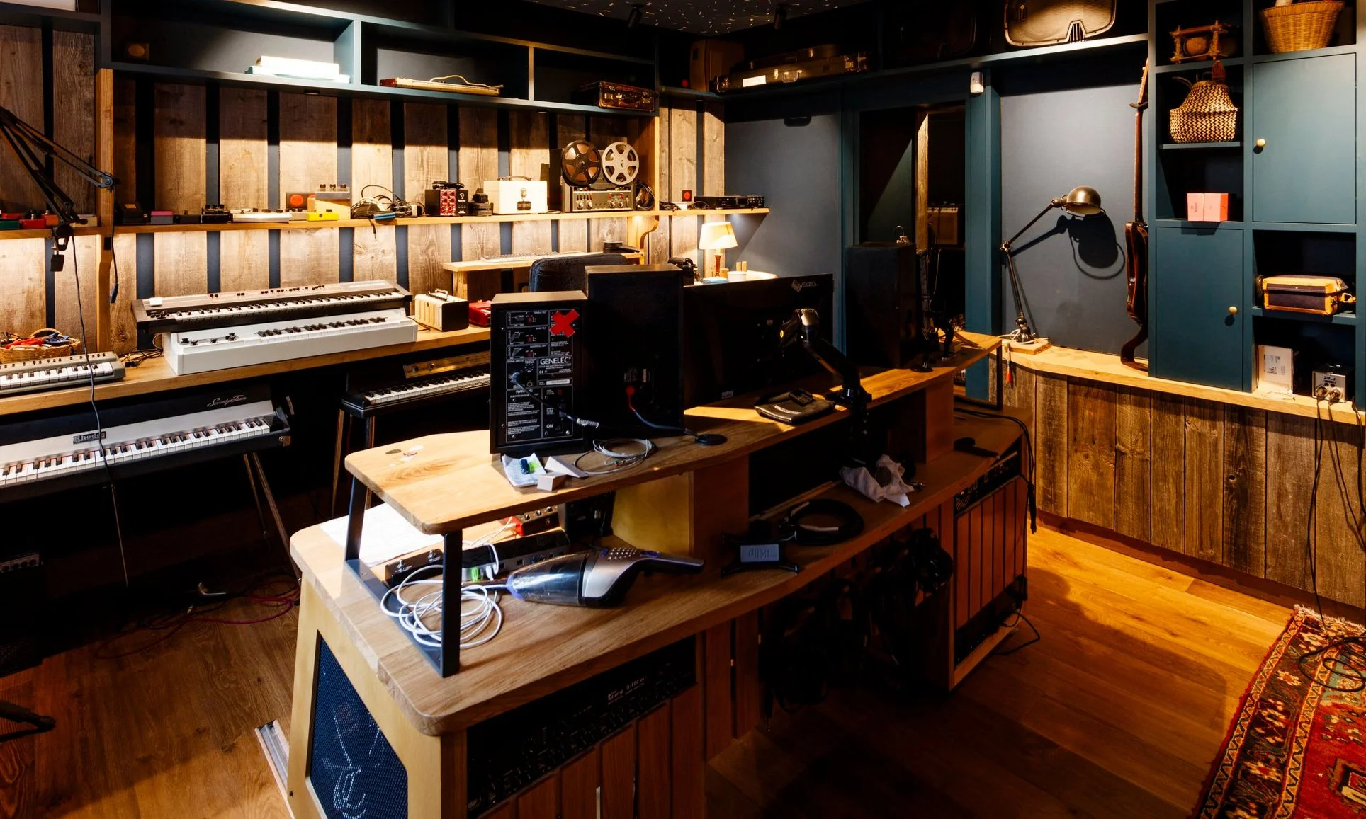 Une pièce équipée d'instruments de musique et d'équipement audio, avec des claviers électroniques, des ordinateurs sur un bureau, et des étagères avec des objets divers, dans un style industriel et chaud.