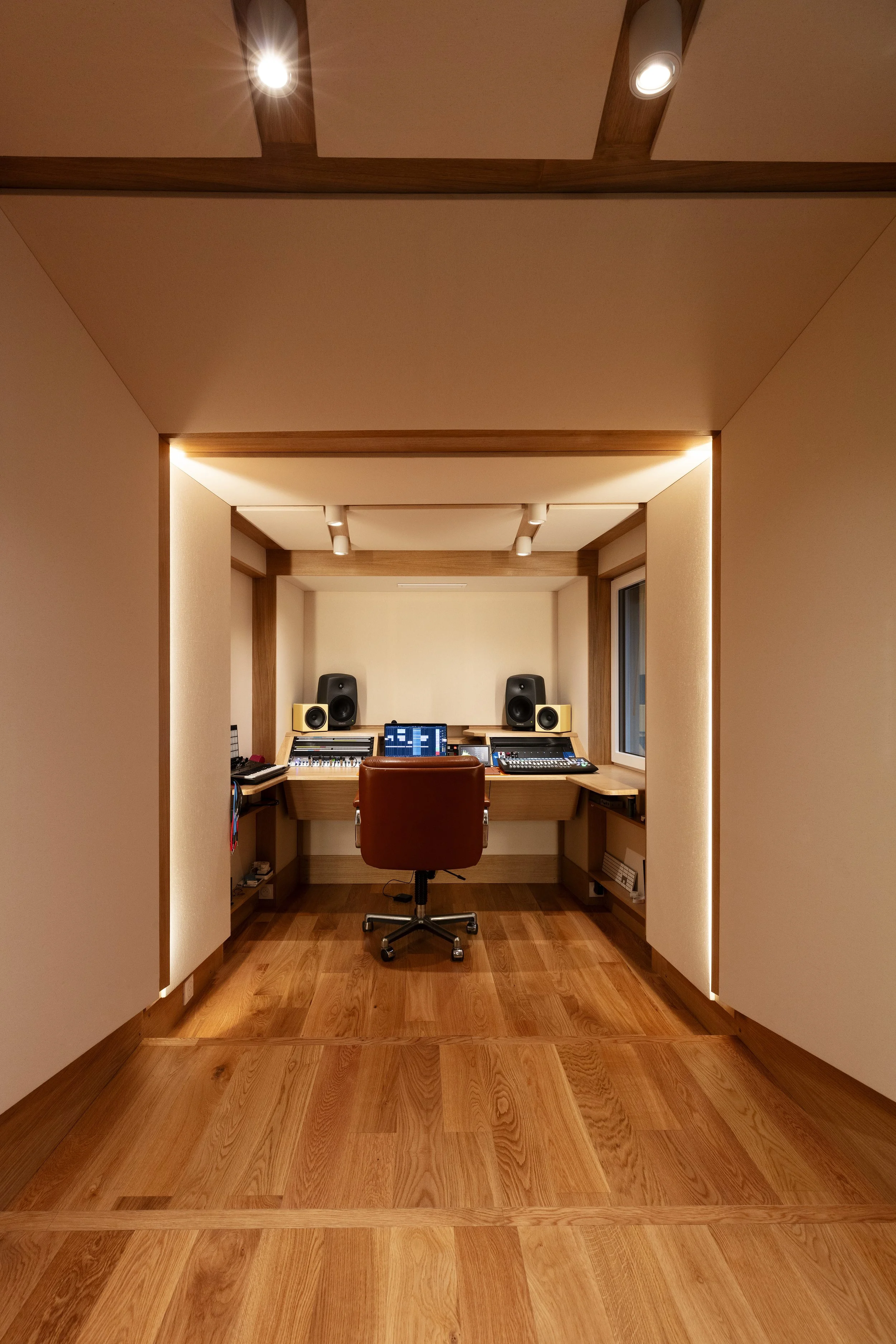 Studio d'enregistrement avec bureau, fauteuil, deux grands haut-parleurs, deux moniteurs de contrôle, et équipement audio, éclairé par lumières encastrées et entouré de murs en bois et panneaux acoustiques.