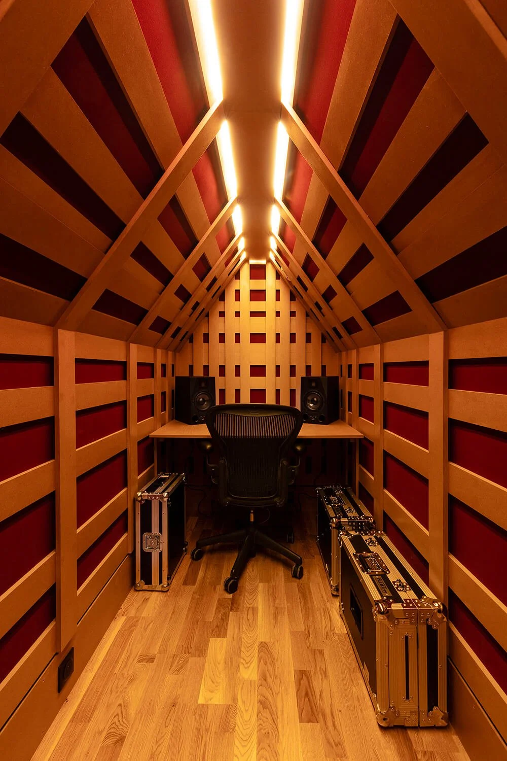Studio d'enregistrement acoustique en bois avec un bureau, une chaise de bureau, deux enceintes, et deux mallettes de matériel technique, ambiance chaleureuse
