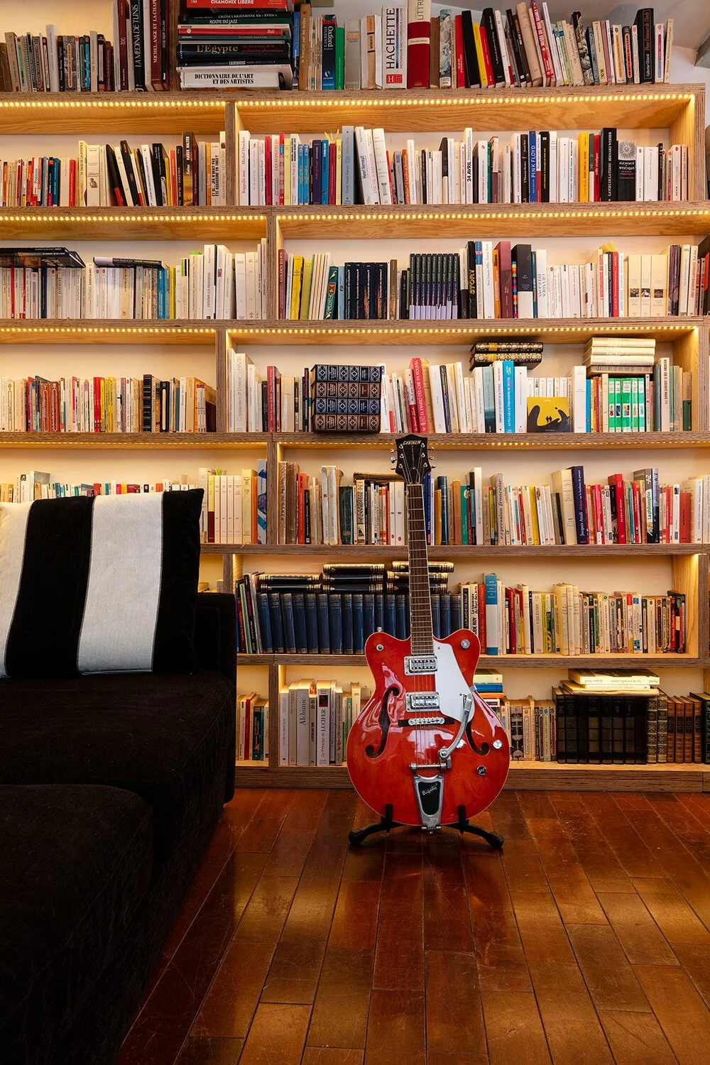 Une bibliothèque avec plusieurs étagères remplies de livres. Au centre, une guitare électrique rouge posée au sol près d'un canapé noir avec un coussin à rayures noires et blanches.
