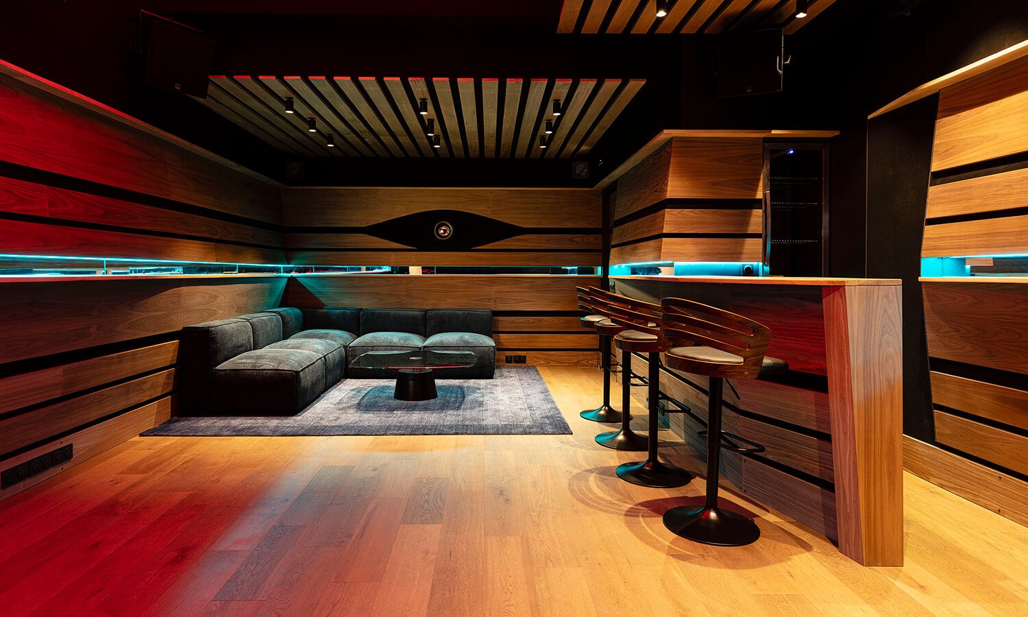 Intérieur d'un lounge ou bar moderne avec un canapé noir, un tapis gris, un plan de travail en bois avec des sièges à bar, murs en bois avec éclairage LED bleu, plafond avec poutres en bois.