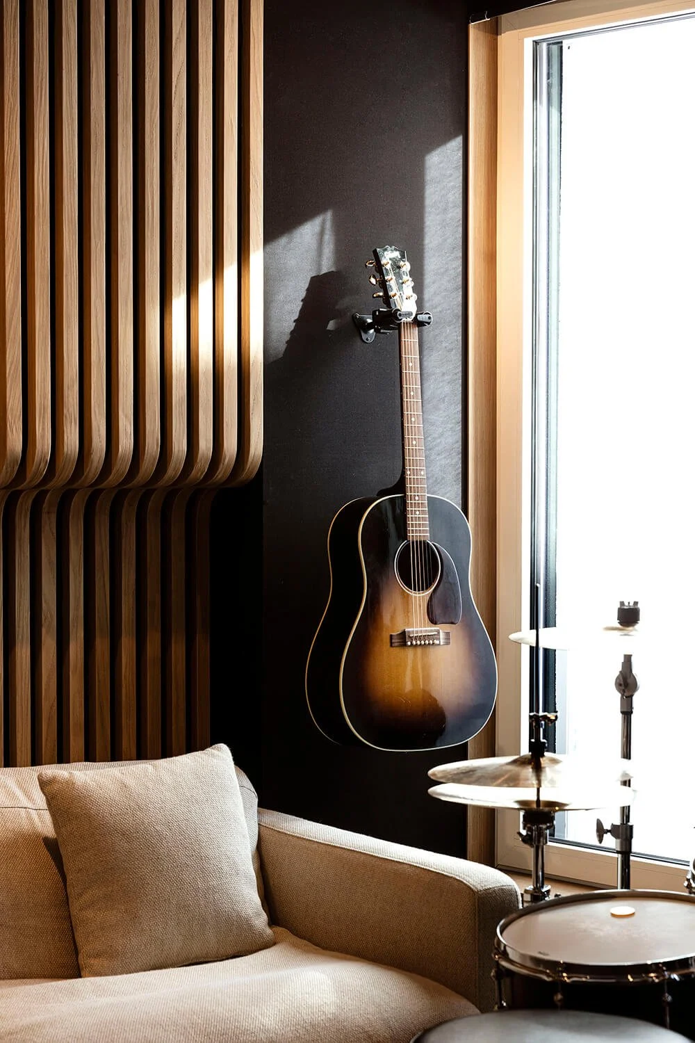 Une guitare acoustique noire suspendue sur un mur dans une pièce moderne, avec un canapé beige en premier plan, et un kit de batterie partiellement visible à droite, près d'une grande fenêtre laissant entrer la lumière naturelle.