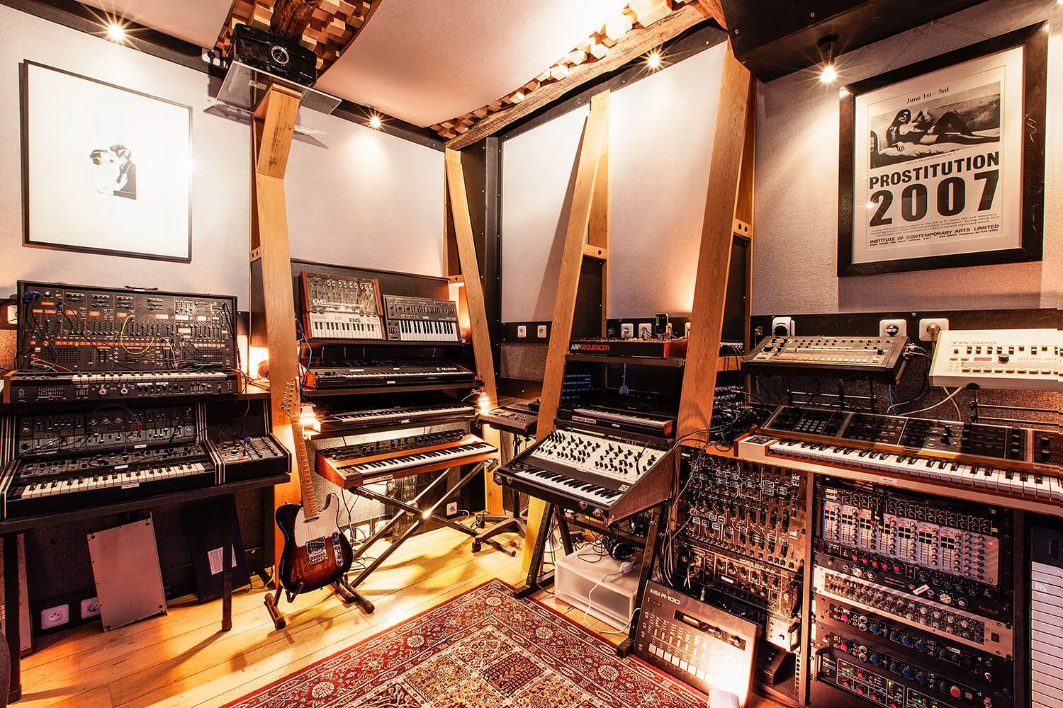 Une salle de musique équipée avec plusieurs synthétiseurs, claviers et équipements audio, décorée avec une affiche de 2007 sur un mur.